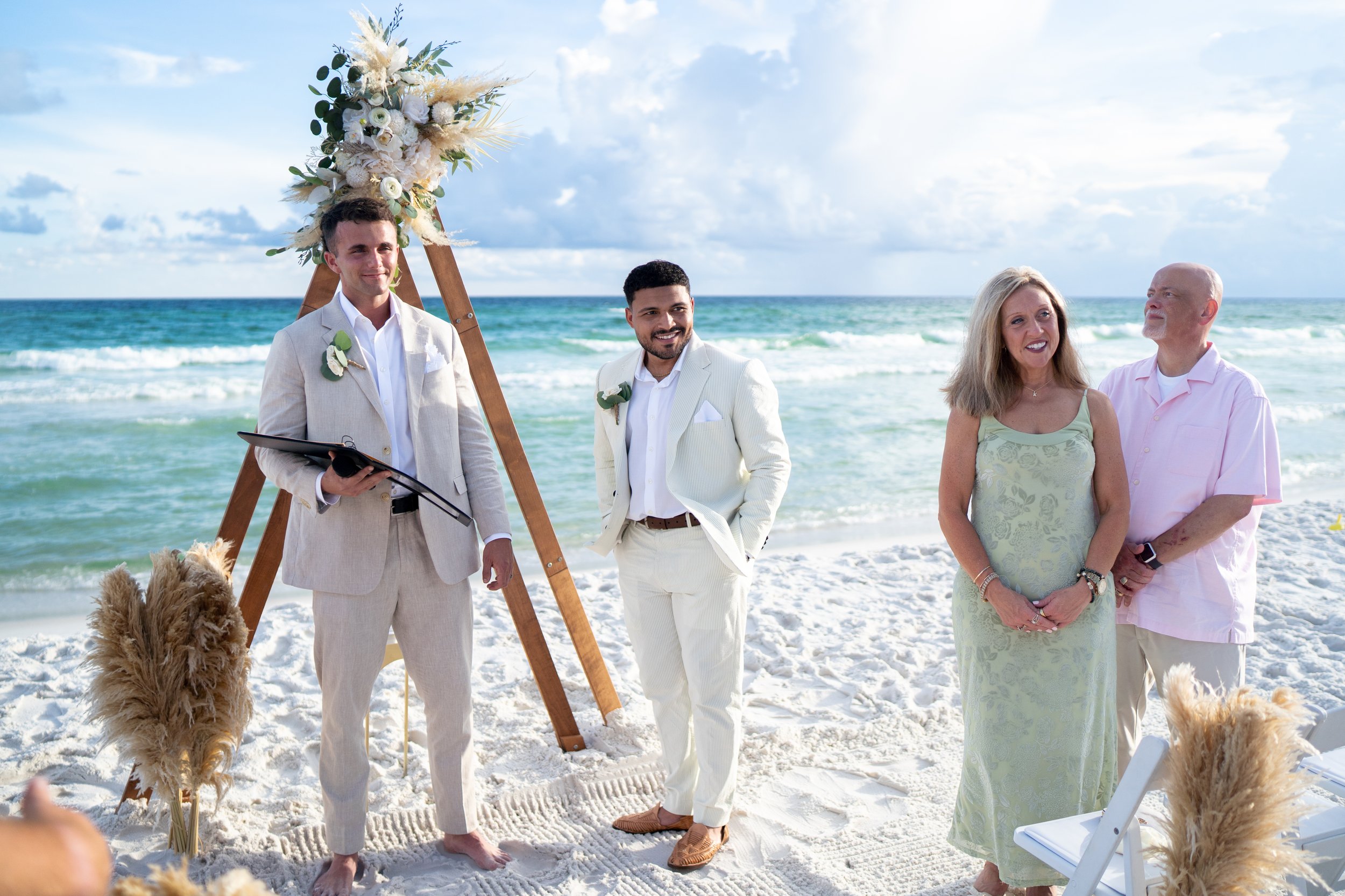 groom and future in laws standing in sandy white sand with the waves of florida's emerald gulf coast behind them as they watch the bride walk down the beach to meet them seaside