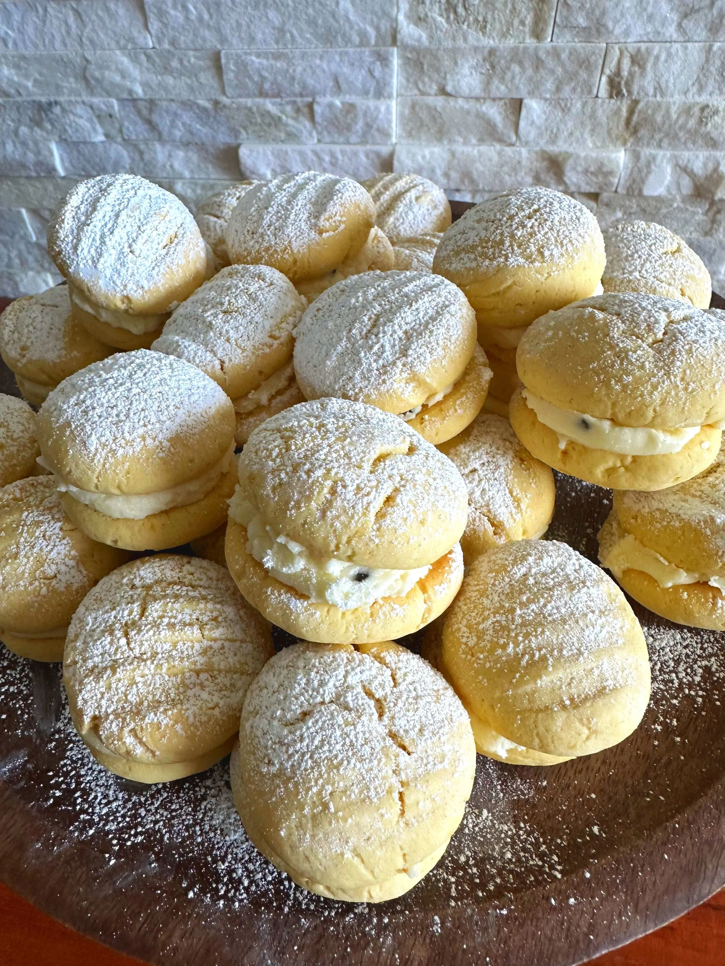 A plate of cream-filled sandwich cookies topped with powdered sugar, arranged on a glass platter against a textured stone wall.