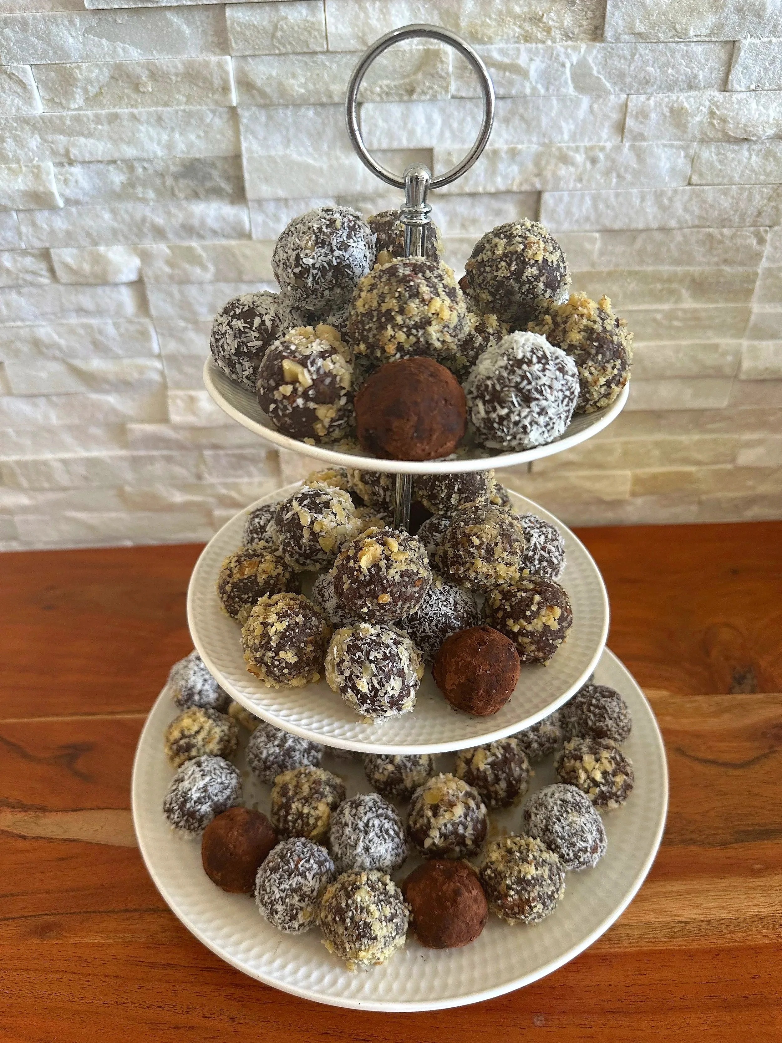 Three-tiered white plate stand filled with assorted chocolate truffles, some covered in coconut, nuts, and cocoa powder, on a wooden surface against a textured white brick wall background.
