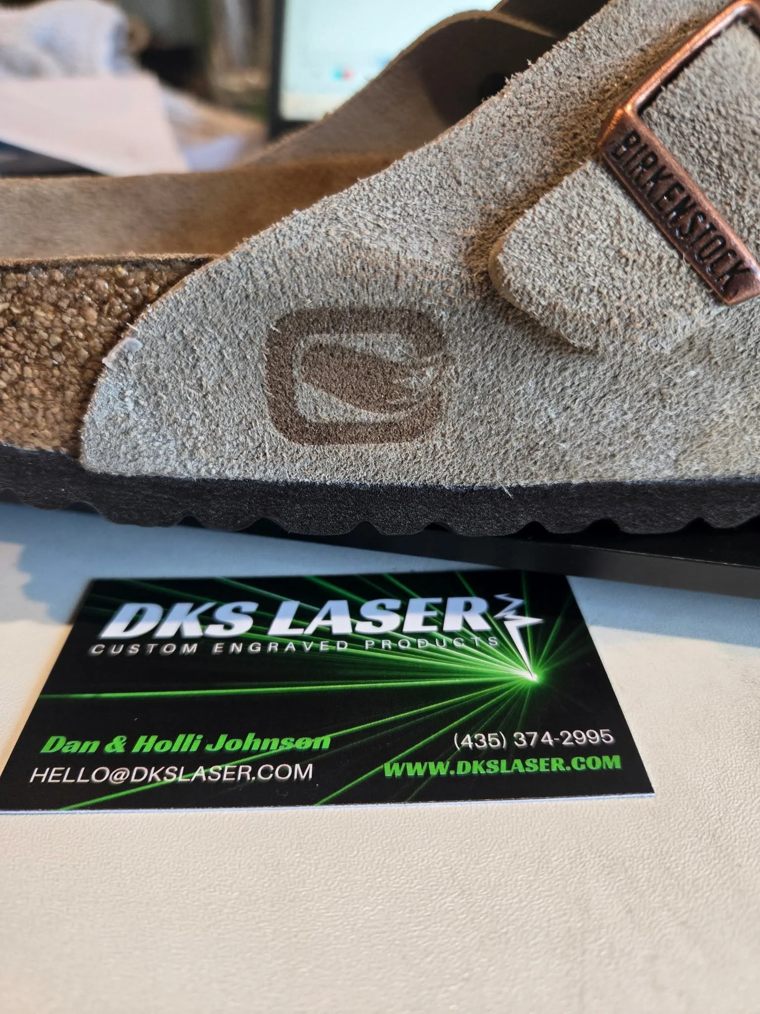 Suede Leather Birkenstock shoe with a rectangular shape logo with a fox tail in the middle of the o that has been laser engraved and burned. The shoe is sitting on a table with a business card of DKS Laser with Dan & Holli Johnson hello@dkslaser.com