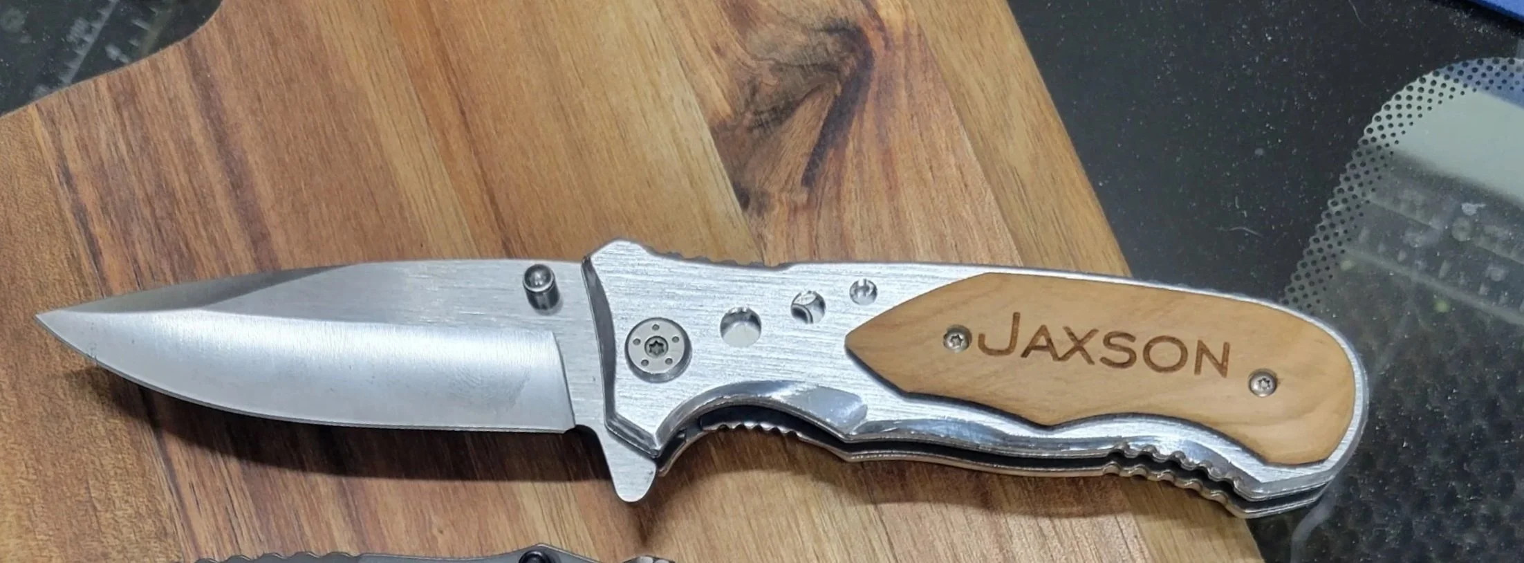 A folding pocket knife with a silver blade and a handle made of wood and metal, engraved with the name 'JAXSON' on the wooden part.