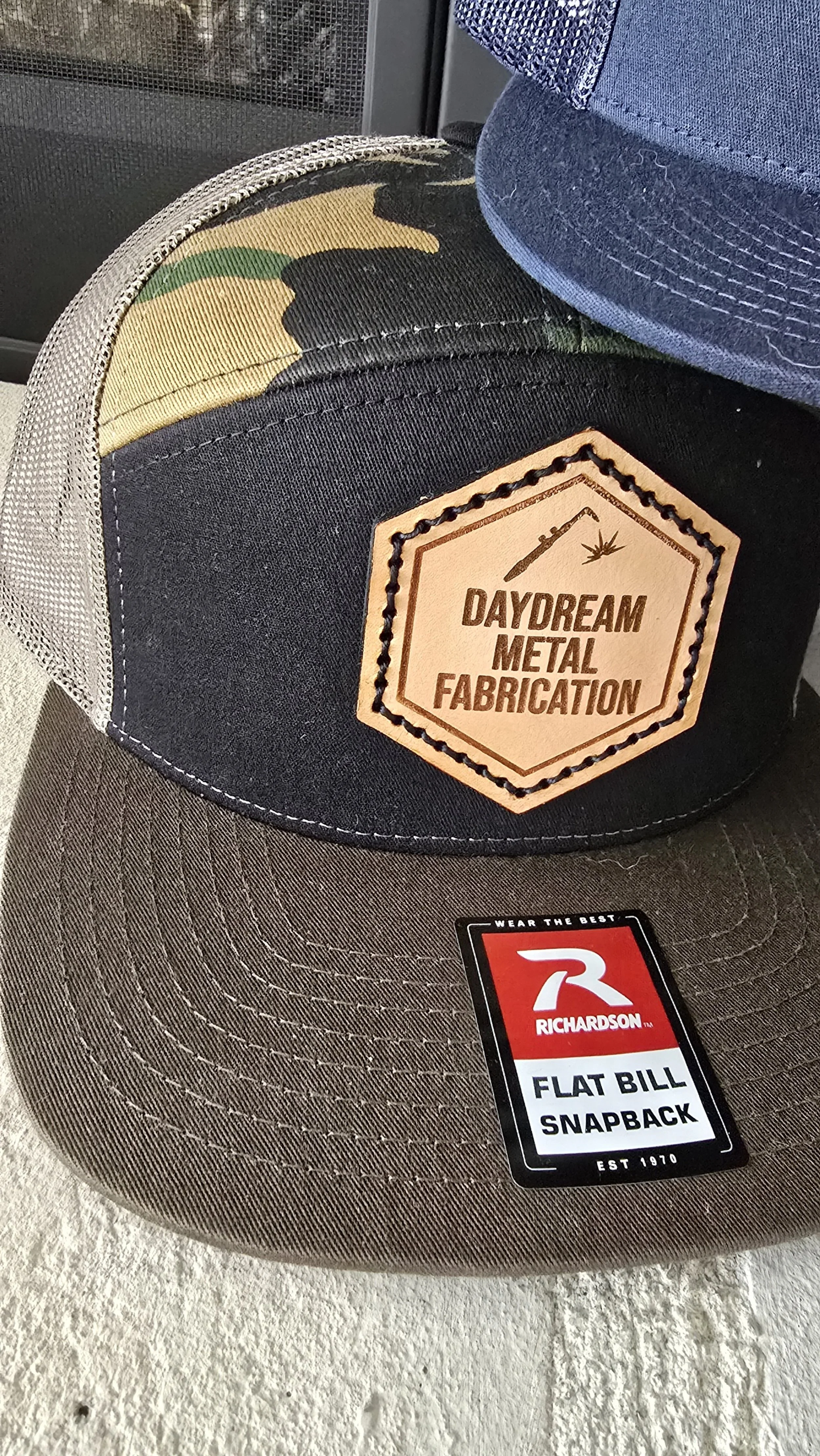 Close-up of a charcoal gray flat bill snapback cap with a hexagon patch reading 'Daydream Metal Fabrication' and symbols of a welding torch and a sticker from Richardson Inc.