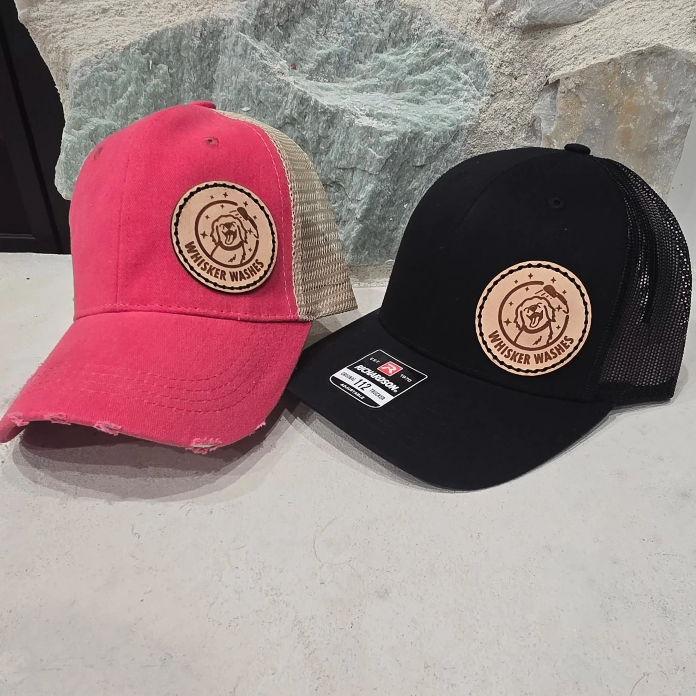 Pink and black caps with a logo reading 'WHISKER WASHES' featuring a dog's face, placed on a light surface with a stone hearth and a dark peek of the fireplace in the background.