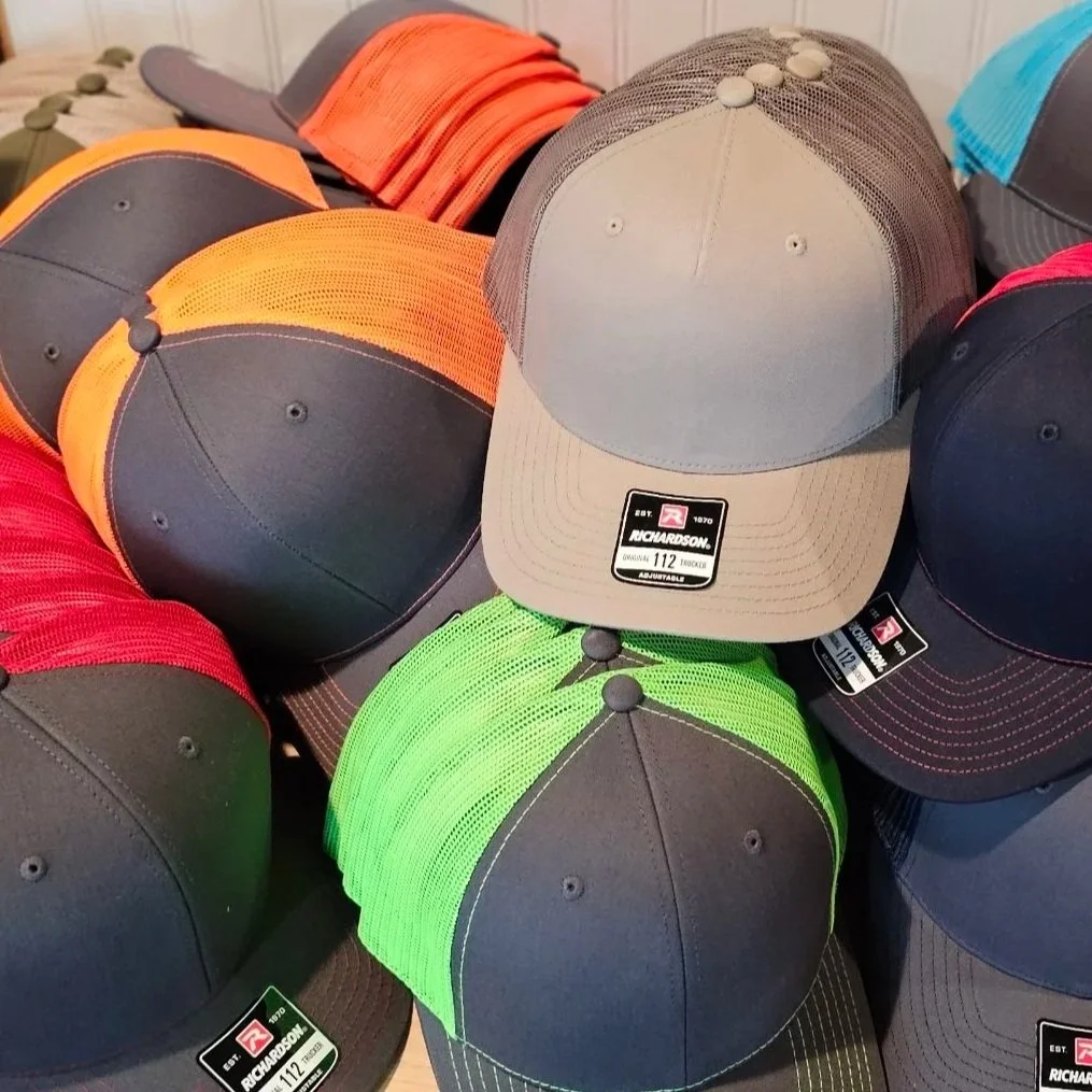 A huge pile of blank Richardson 112 hats with multiple color options orange/charcoal, charcoal/red, neon green/charcoal, neon blue/charcoal and then a light grey/charcoal color.