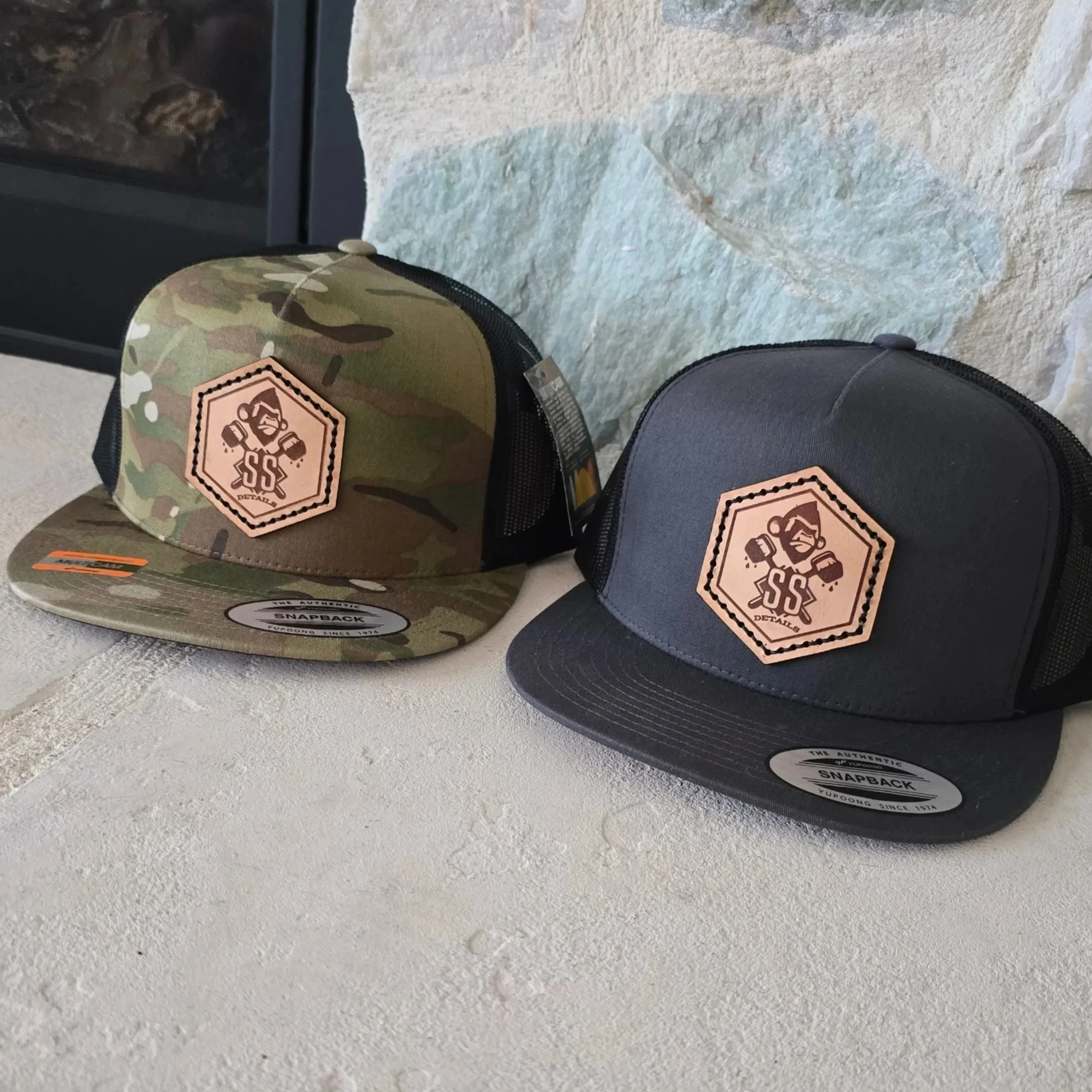 2 different hats with a leather patch hat sewn on to a camoflauge and grey/charcoal flat bring Yupoong 6006 Classic Trucker Cap. The patch features a gorilla holding some brushes with SS and details under it. The hats are sitting on a rock hearth.