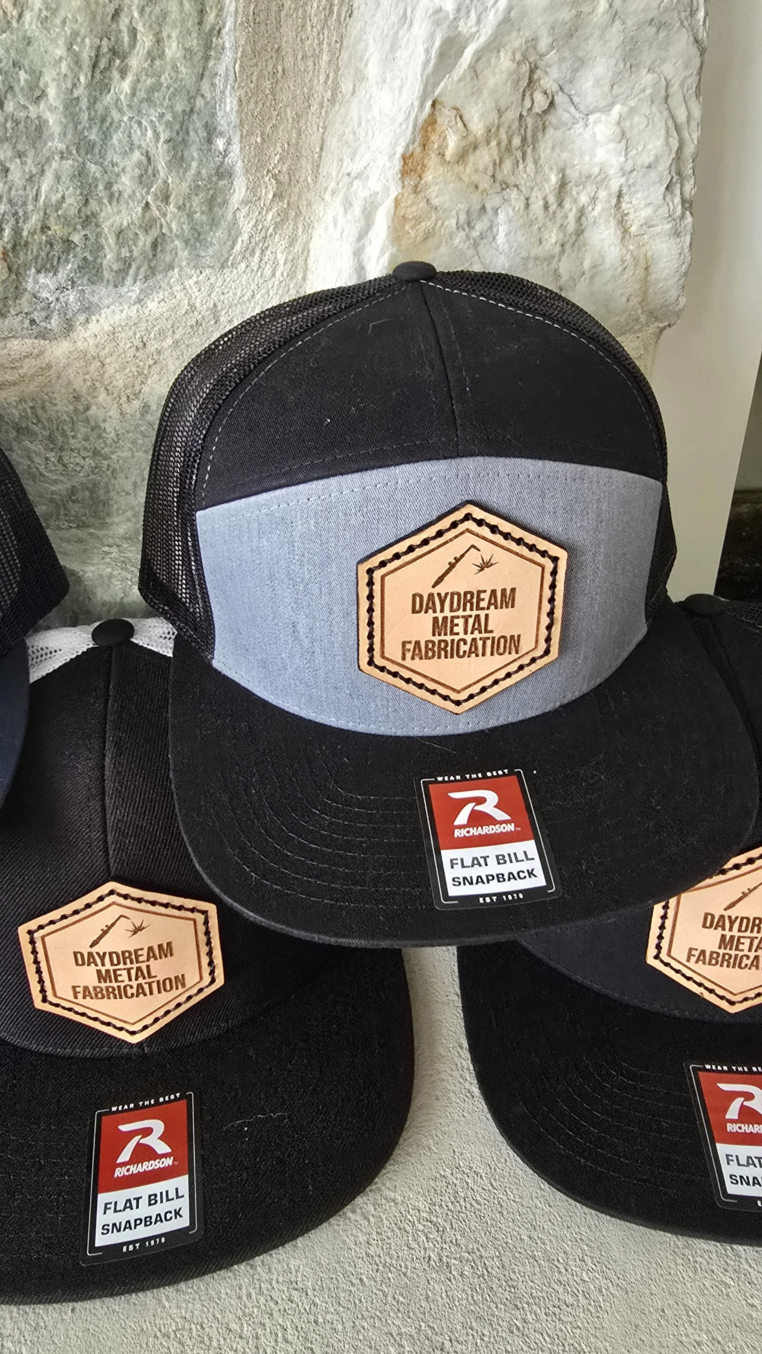 Three black and gray snapback hats with labels  "Richardson Flat Bill Snapback."  one has a patch that reads "Daydream Metal Fabrication" with a welding torch.