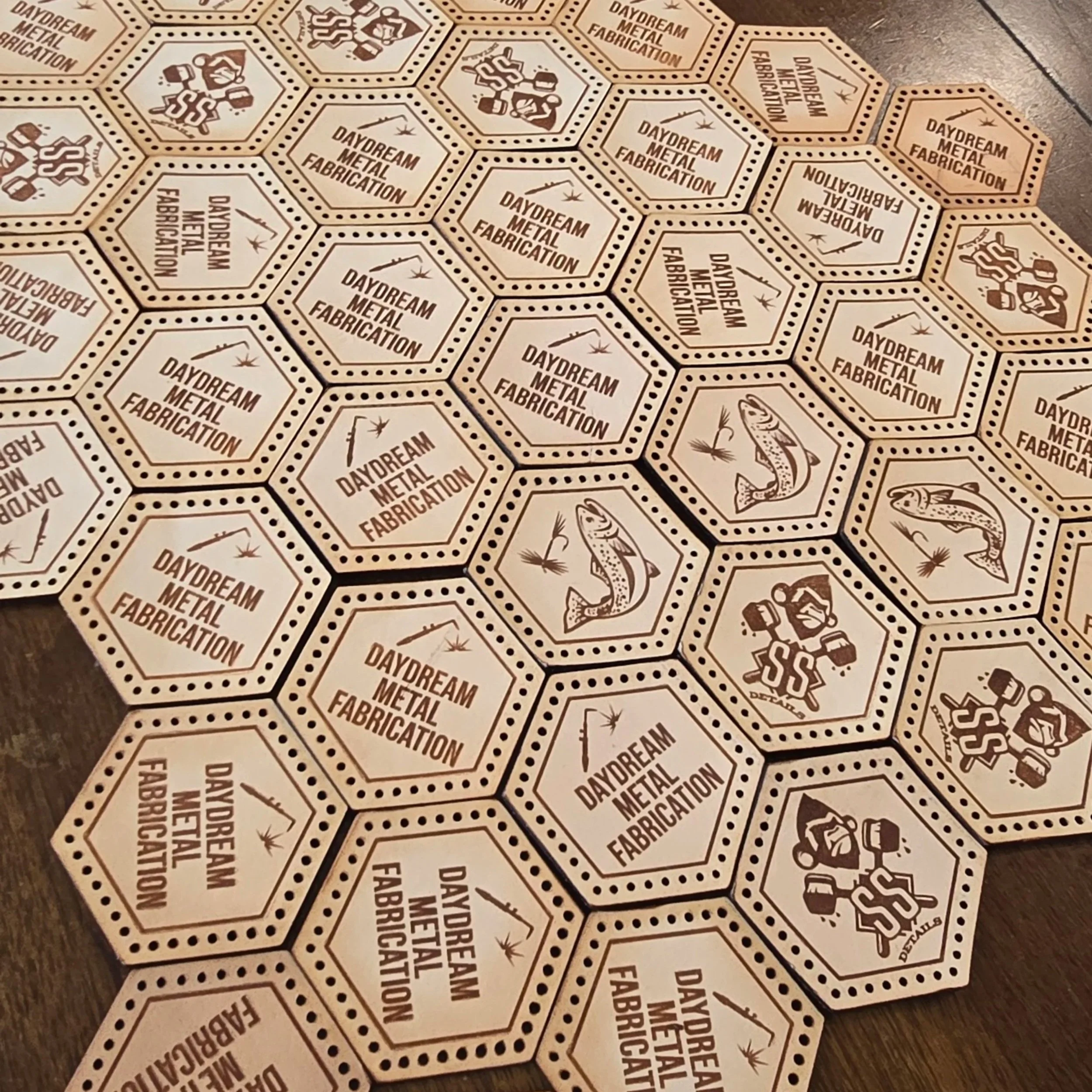 A lot of leather patches in hexagon shapes that look like honey comb or soccer ball pattern in how they are spread upon the table. They have various logos on them for Daydream Metal Fabrication, SS Details and a trout fish.
