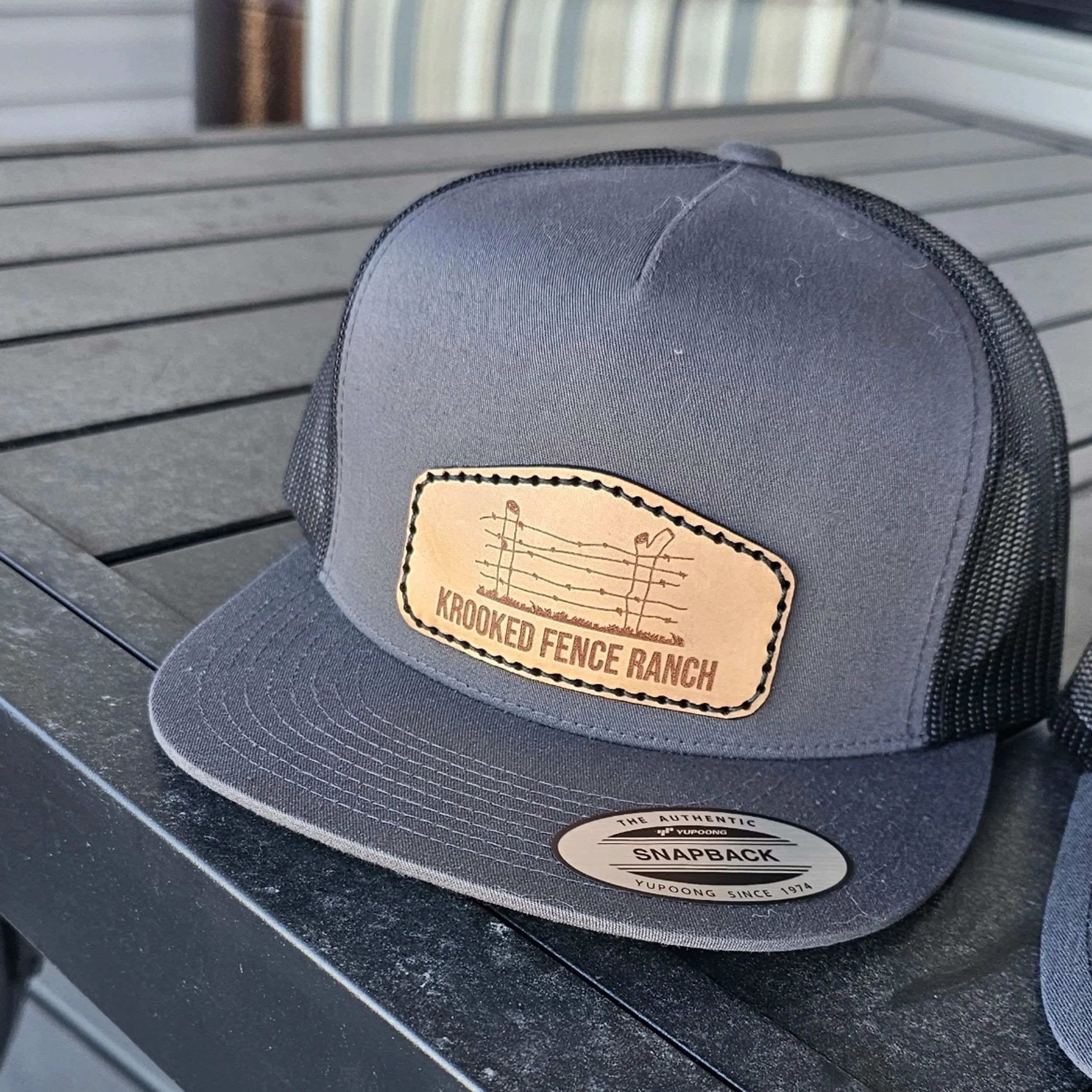 Gray snapback hat with a leather patch displaying a barbed wire fence and the text "Krooked Fence Ranch" on a black outdoor table.