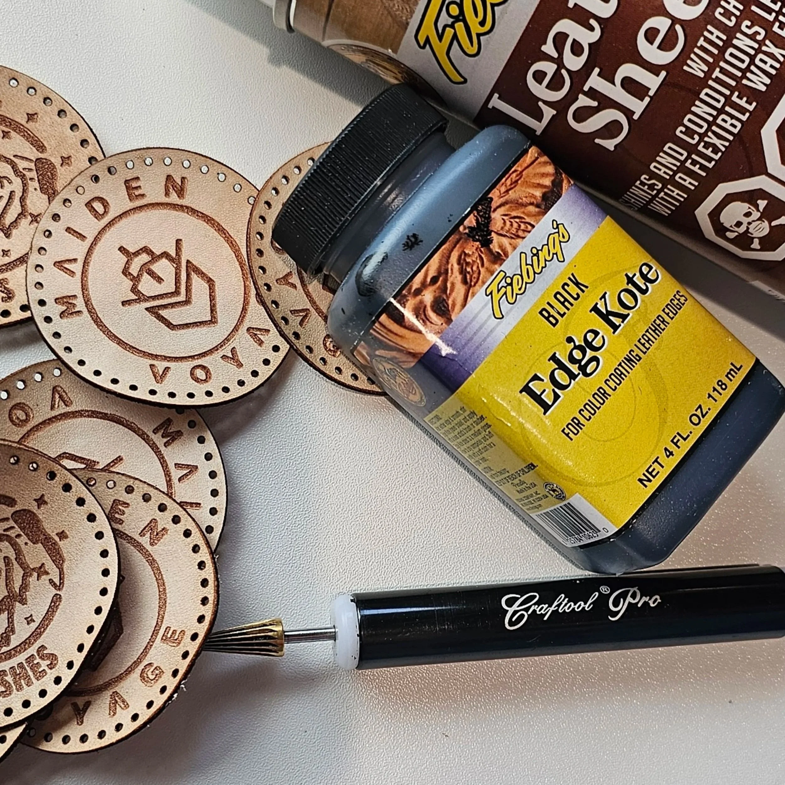 A few round shaped leather patches with Maiden Voyage logo layered with Edge Coat, Leather Sheen Spray and a Leather working tool.