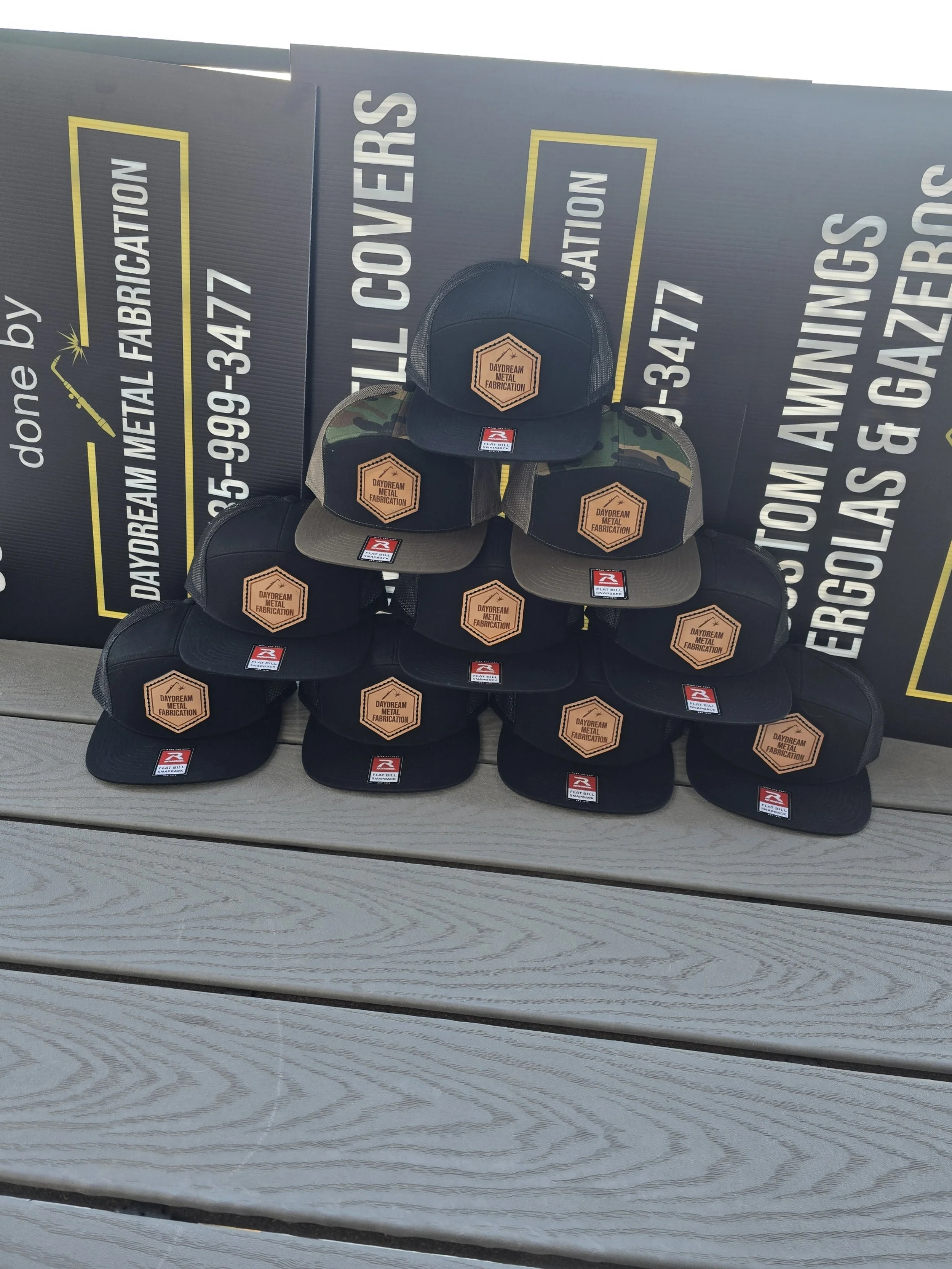 Black & Camo 7 panel Richardson Hat with a hexagonal shape leather patch sewn on the center of the hat. 10 hats stacked in a pyramid on a grey wood looking floor.