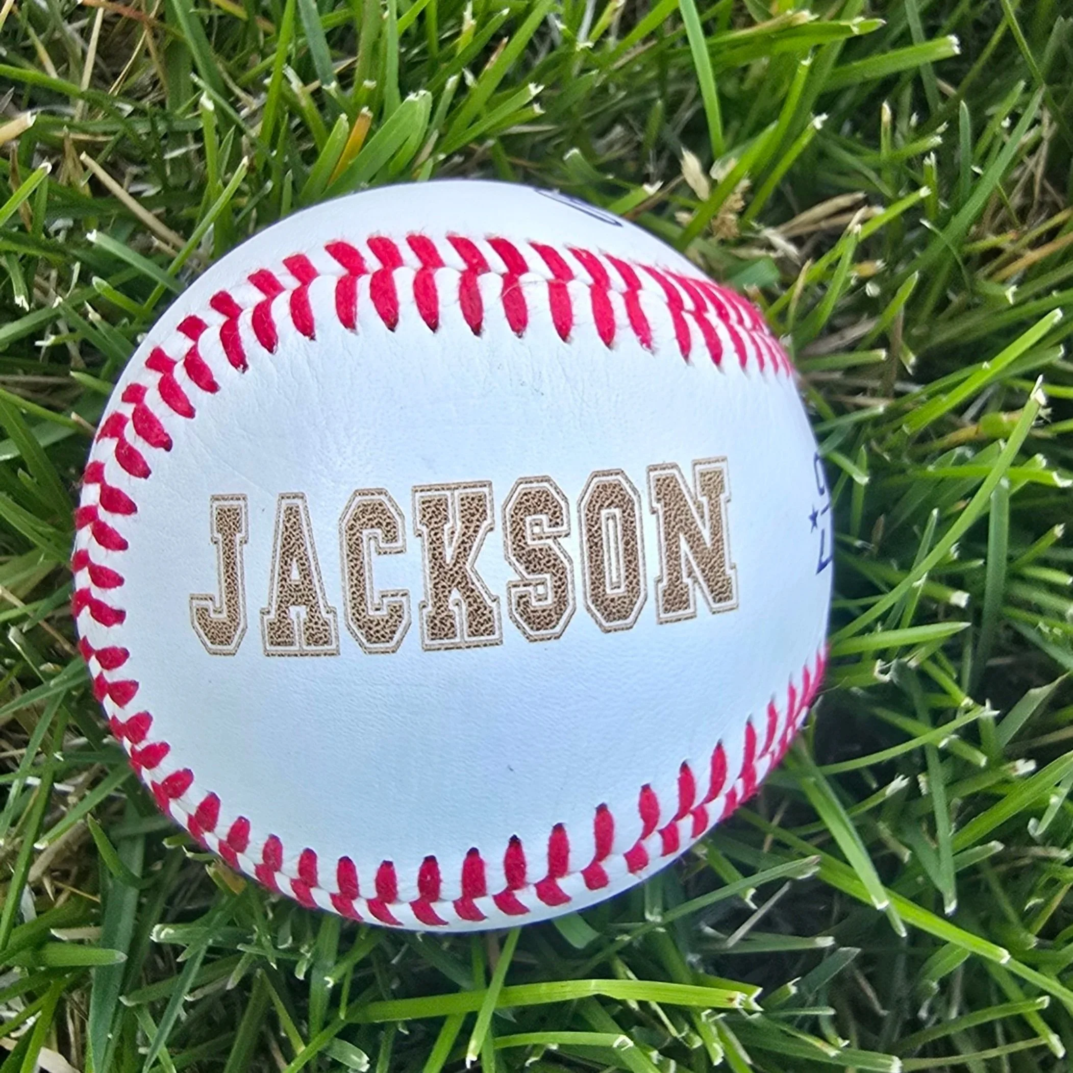 Laser Engraved "JACKSON" on a white traditional baseball that is laying in the grass.