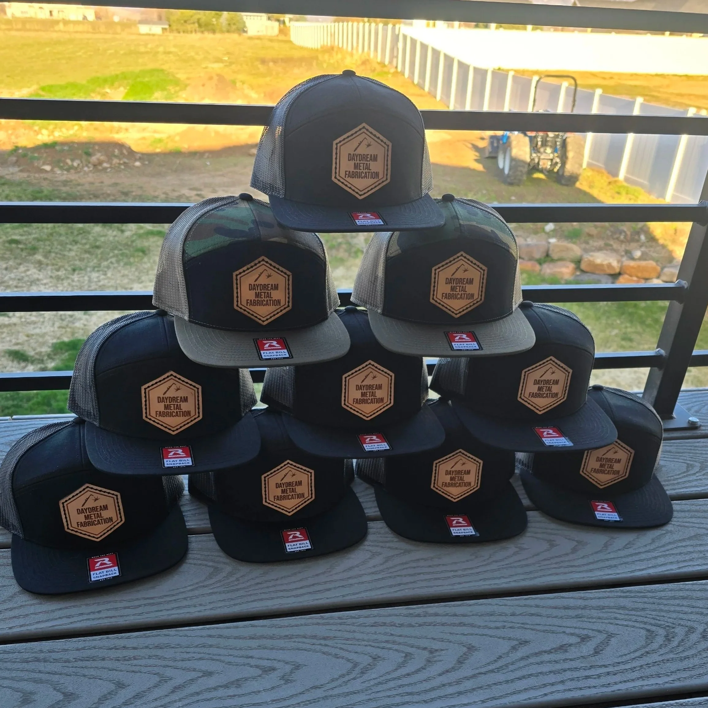 Stack of ten black and gray trucker hats stacked up like a pyramid with leather patches that say 'Daydream Metal Fabrication' on a deck, with a backyard and fence in the background.