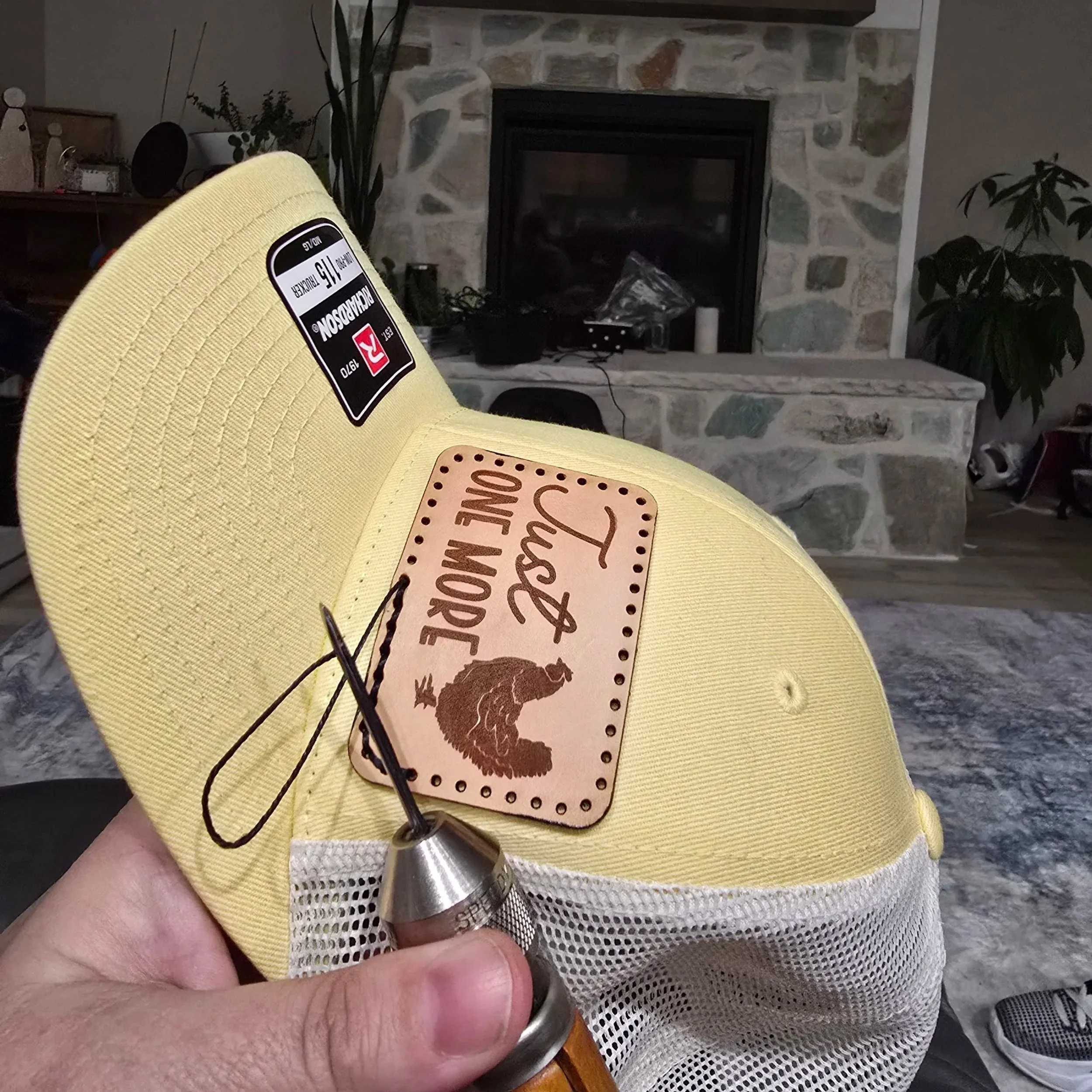 Person hand stitching a rectangle leather patch with "Just One More and a chicken" laser engraved on to a leather patch. Hand sitching it on to a Richardson 115 hat style while sitting in front of a cozy fireplace.