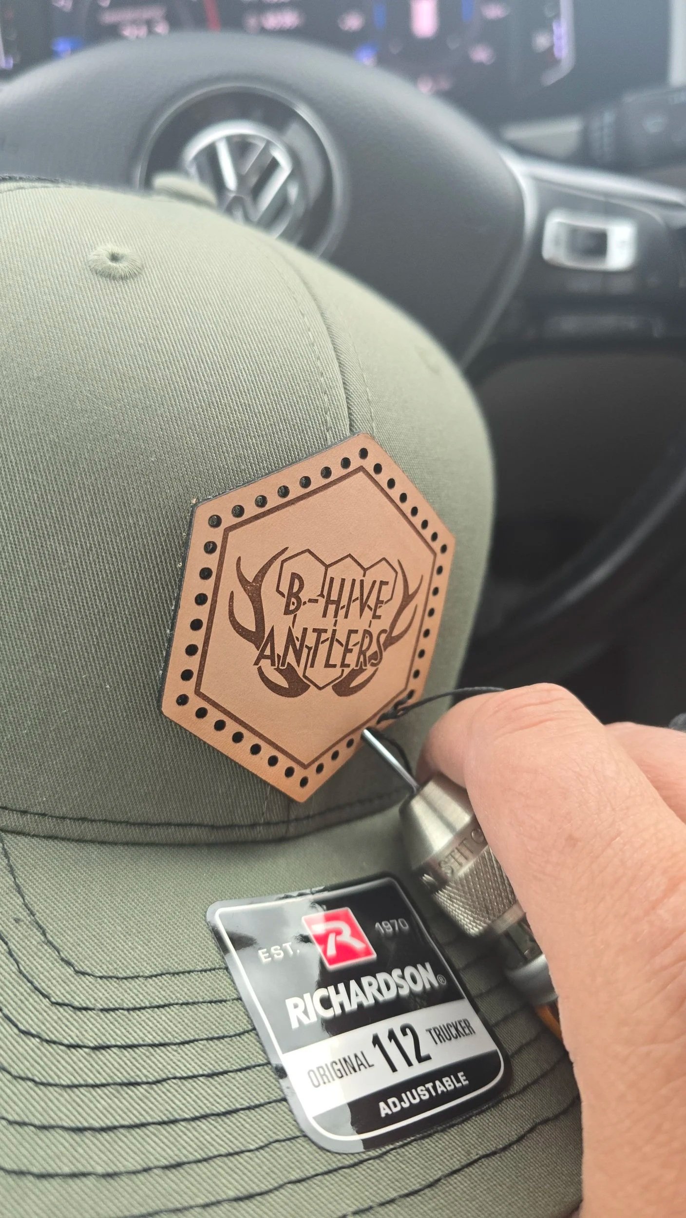 An olive green hat with a hexagonal shape leather patch with holes & dark inner outline matching the exterior shape. Two enclosed antlers with honeycomb shapes with B - Hive Antlers in all capitals overlaying. Being hand stitched on with a needle awl