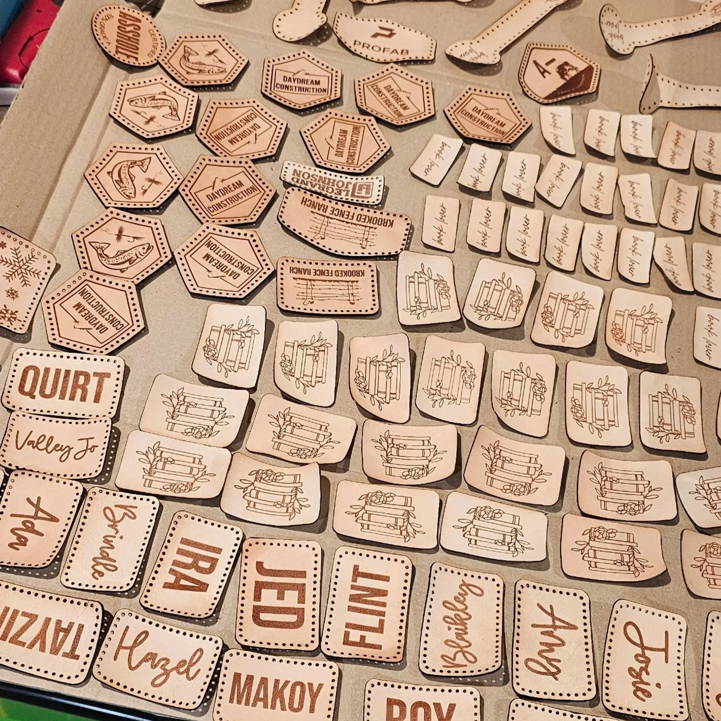 A spread of different shaped and sized leather patches laying on a table. Some have logos, some have books, fish, and others are filled with names in various fonts and sizes.