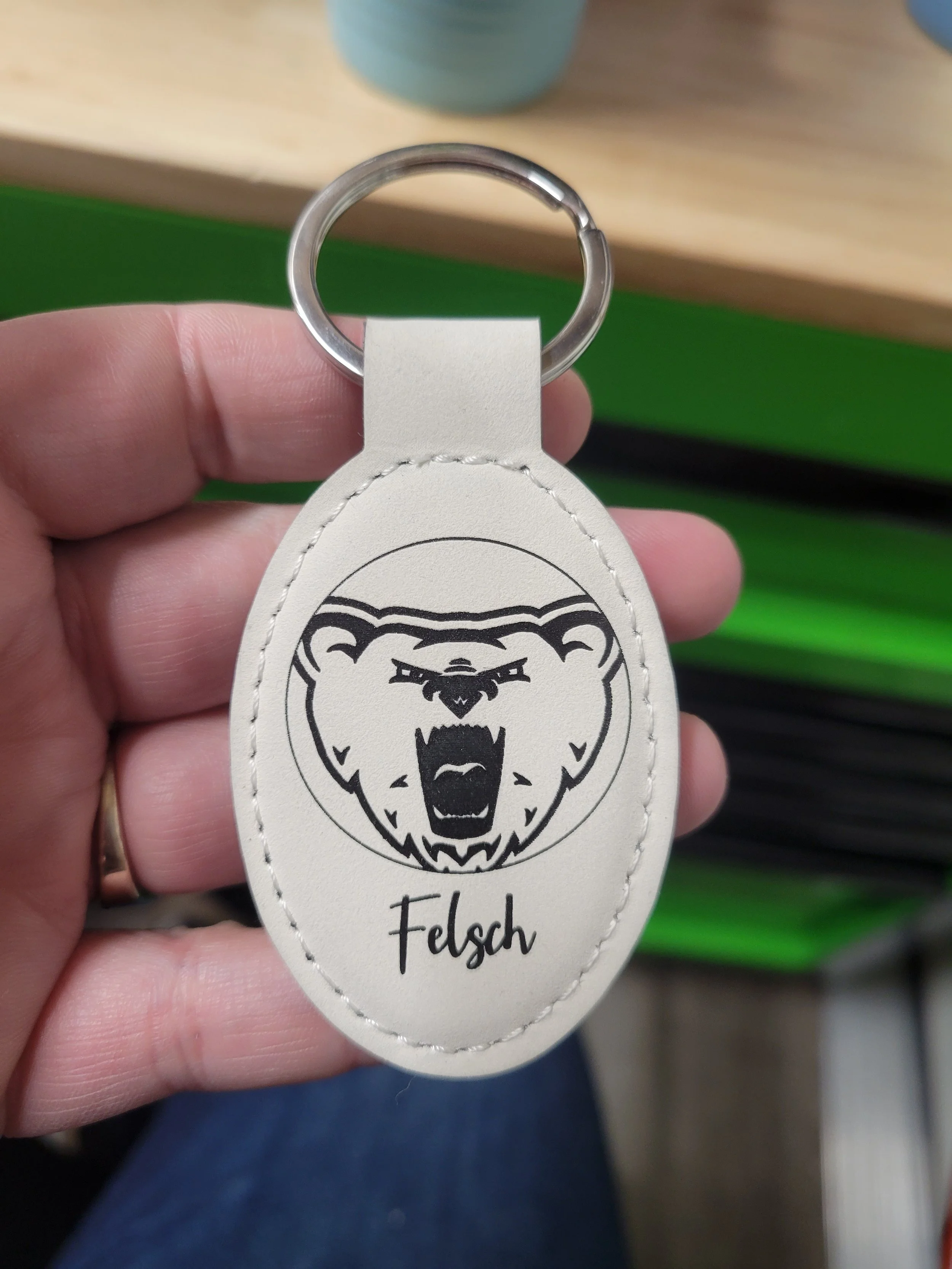 A white, oval keychain with a black laser engraving of a bear face growling inside of a thin line circle. The name Felsch written below it