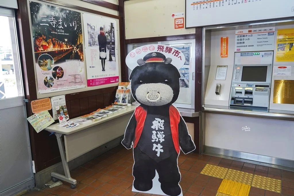 Inside a Japanese train station with posters, brochures, and a black bear mascot cutout near a ticket machine.