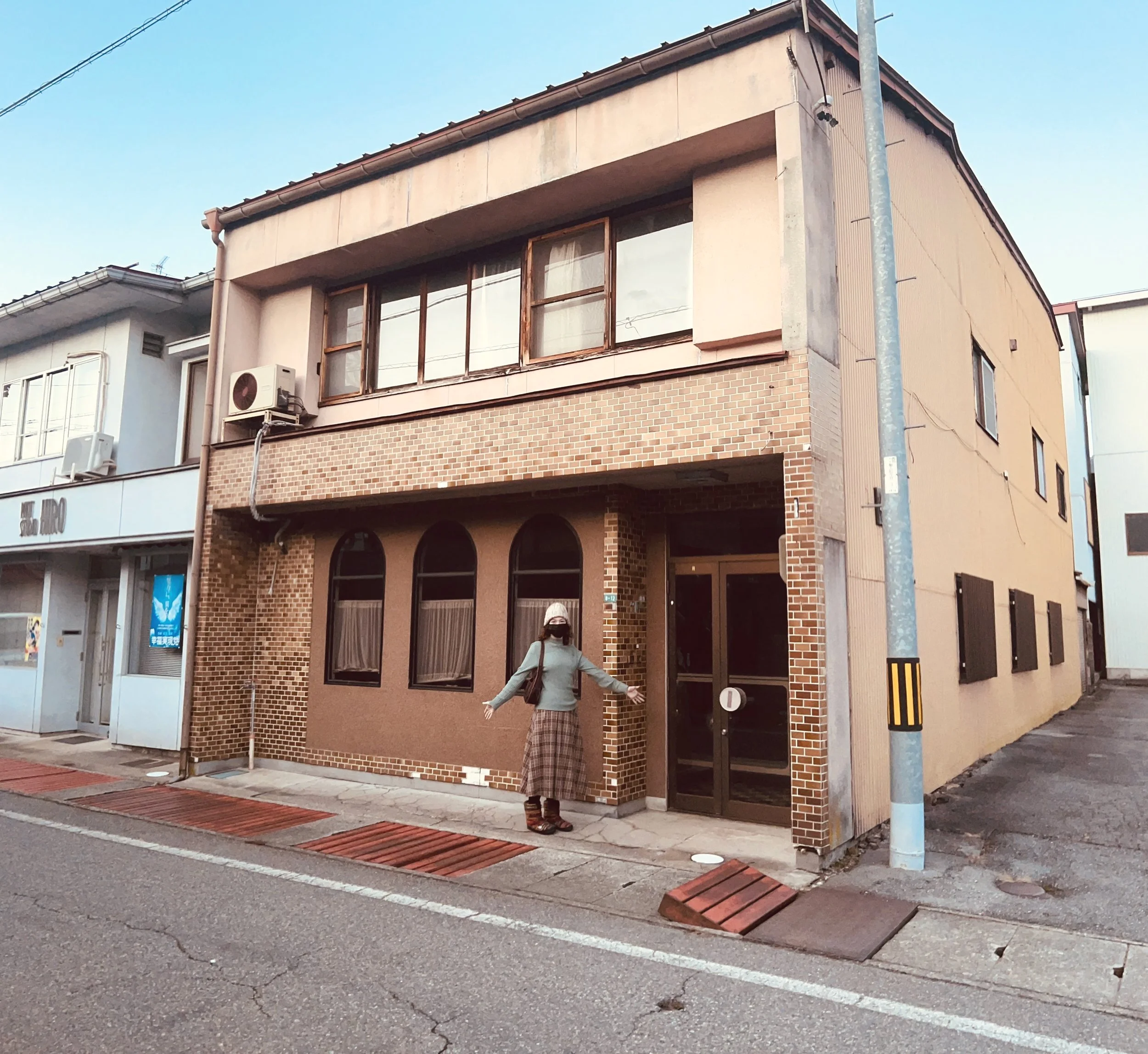 Person standing in front of a two-story building with a brick facade and large windows on an urban street. The building has a flat roof and adjacent is a smaller structure with signage. The street is empty with visible sidewalks and road markers.