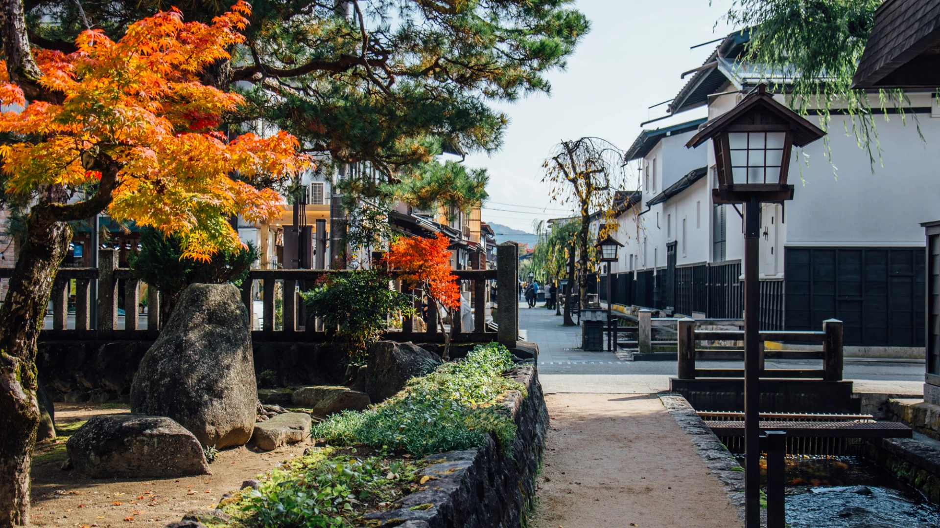 Japanese street scene with traditional architecture, trees with autumn foliage, stone pathway, and wooden lanterns.