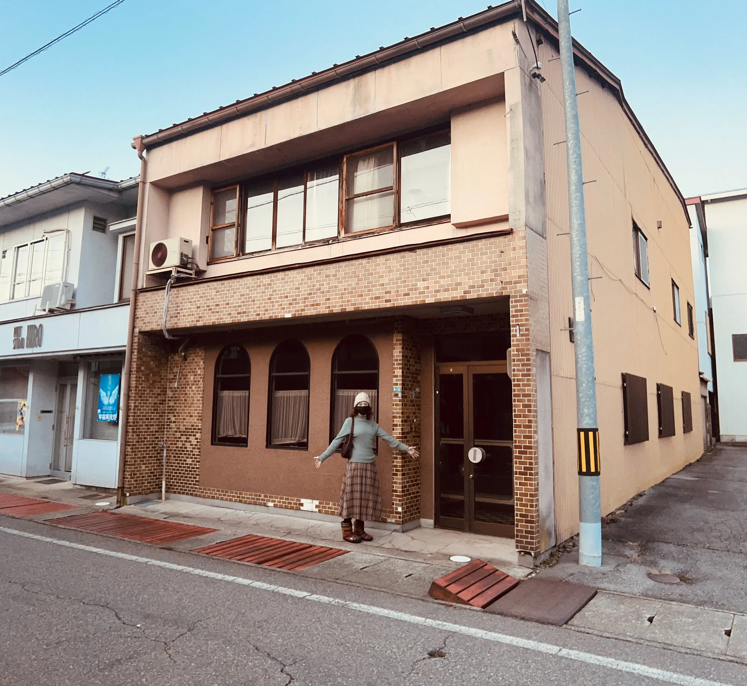 A person standing outside a two-story building with a brick façade in a street setting. The person is wearing a mask, a cap, and a long skirt. The building has large, arched windows and an air conditioning unit on the wall.