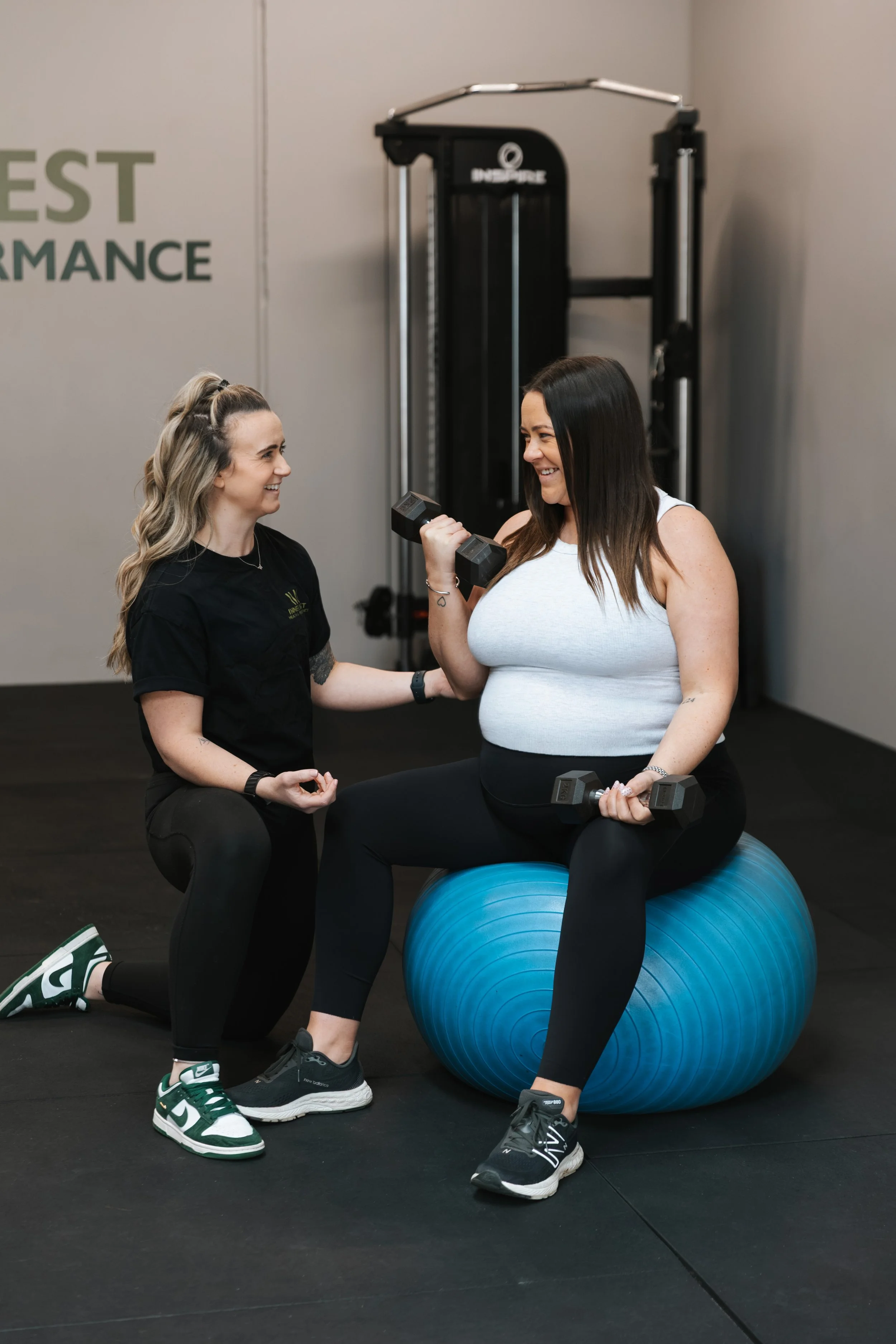 Kirby, with a pregnant client sitting on an exercise ball performing a dumbbell bicep curl with Kirby supervising. they are smiling at each other