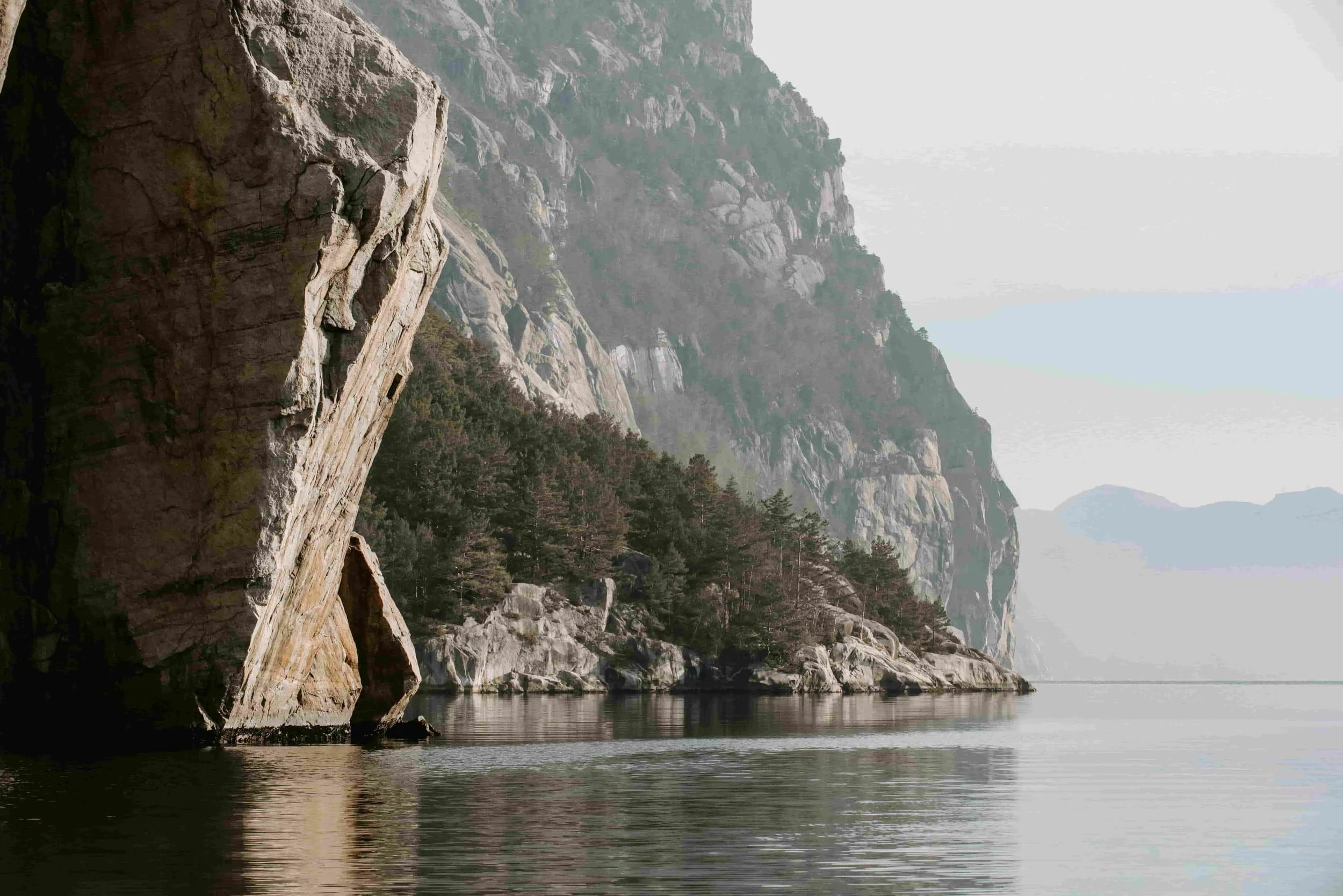 Steiner og trær ved en rolig innsjø i et fjellandskap, med tåkete fjell i bakgrunnen.