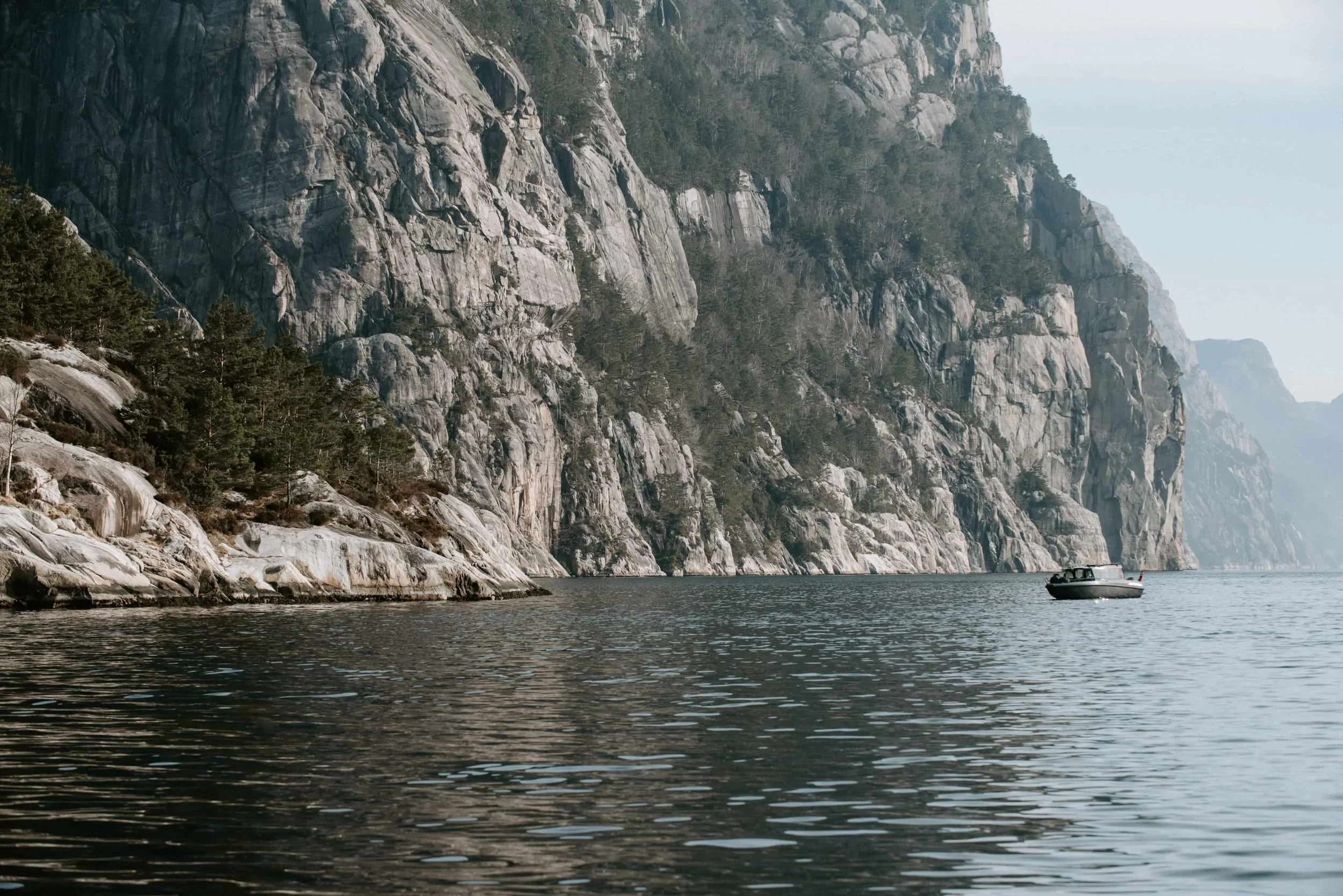 Ein Boot fährt auf einem See entlang einer felsigen Steilküste mit Bäumen an der Spitze.