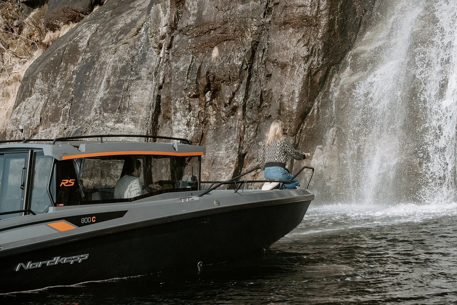 Én person sitter i baugen på en båt som seiler på en elv, mens en annen person spruter vann fra en fjellvegg.