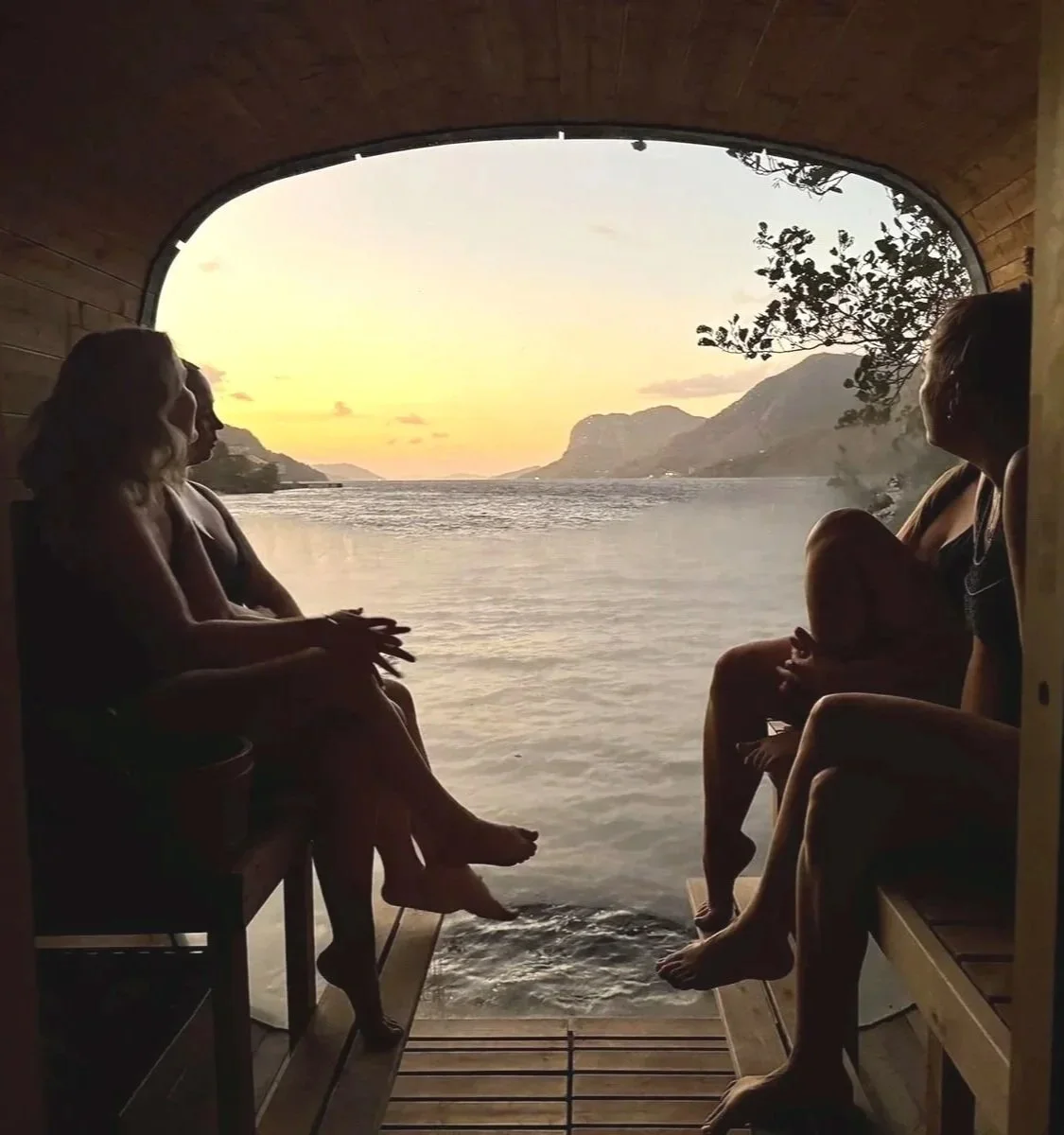 Drei Frauen sitzen in einer Sauna mit Blick auf einen See bei Sonnenuntergang, umgeben von Bergen.