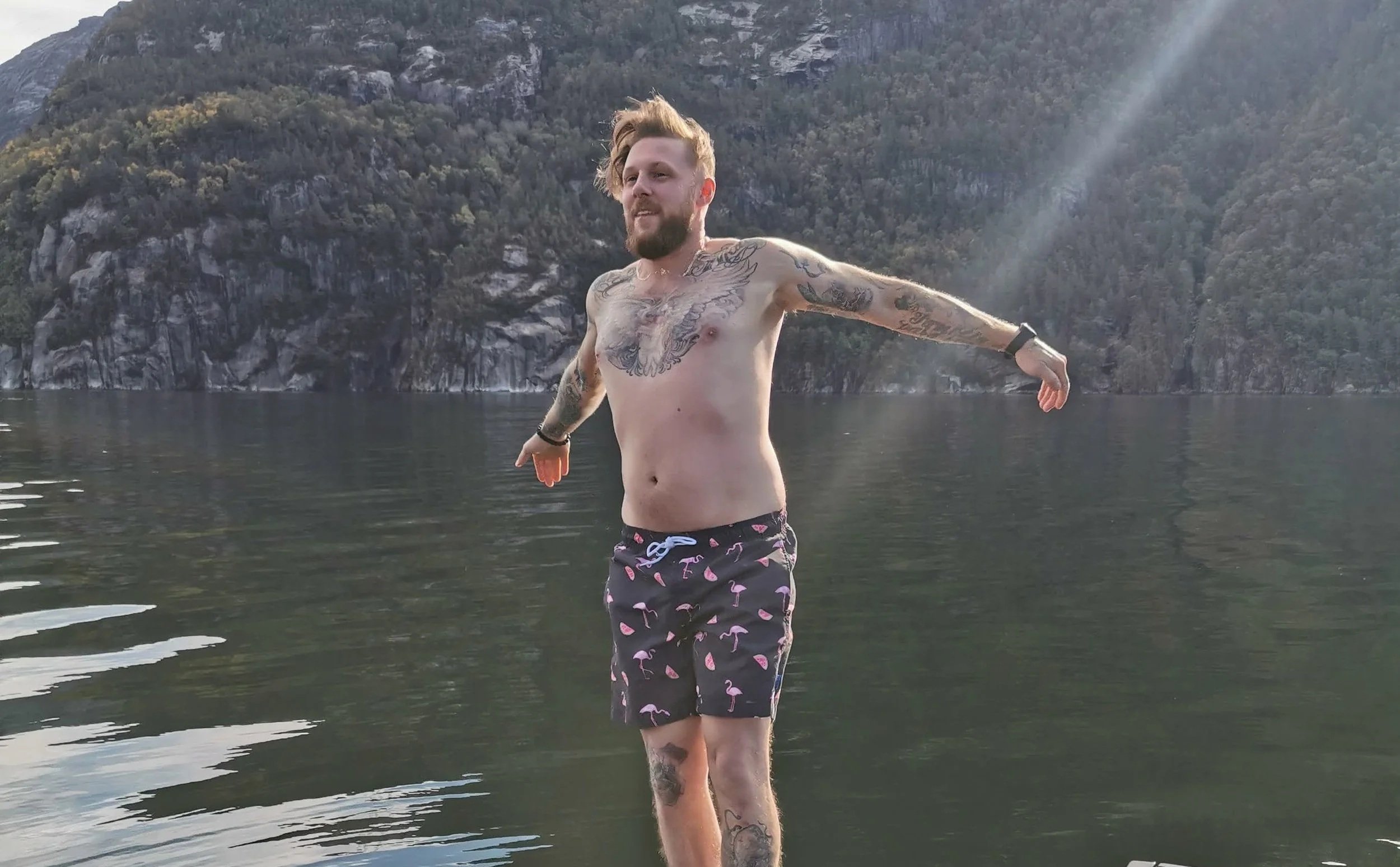 Ein Tattoo-Model steht nackt im Wasser eines Sees, mit Bergformationen im Hintergrund, Arme ausgestreckt, Sonnenstrahlen scheinen auf ihn.