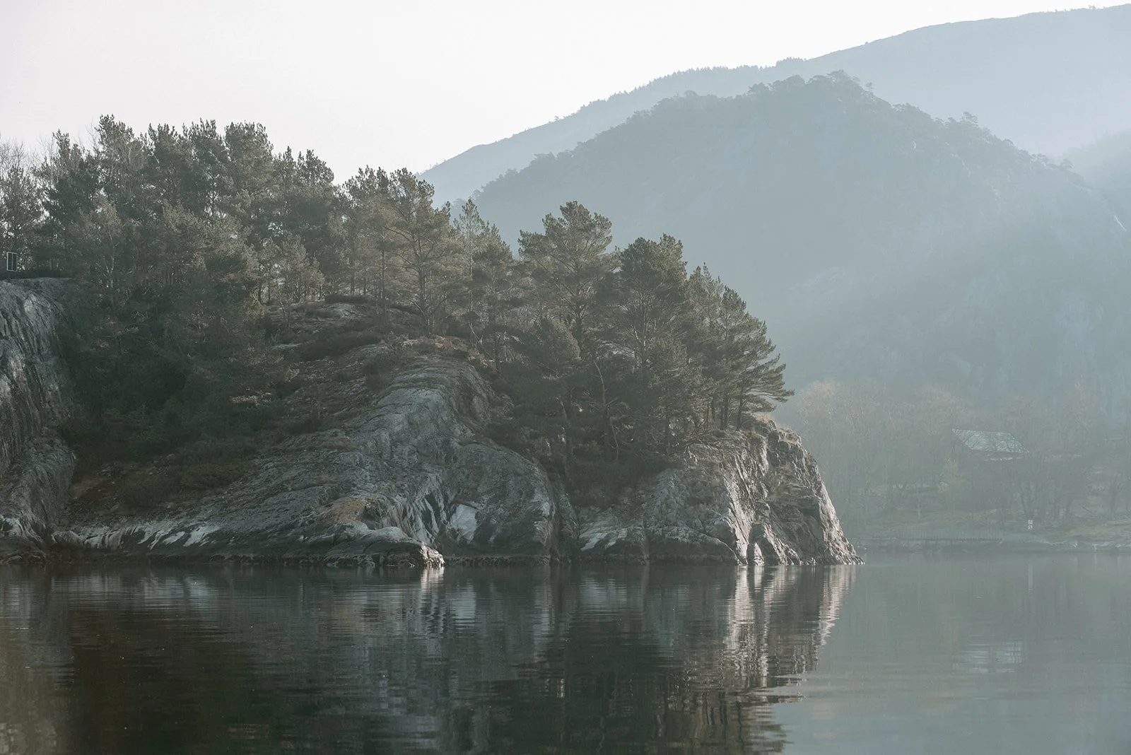 Et tåkete fjellandskap med steiner, trær og rolig vann.