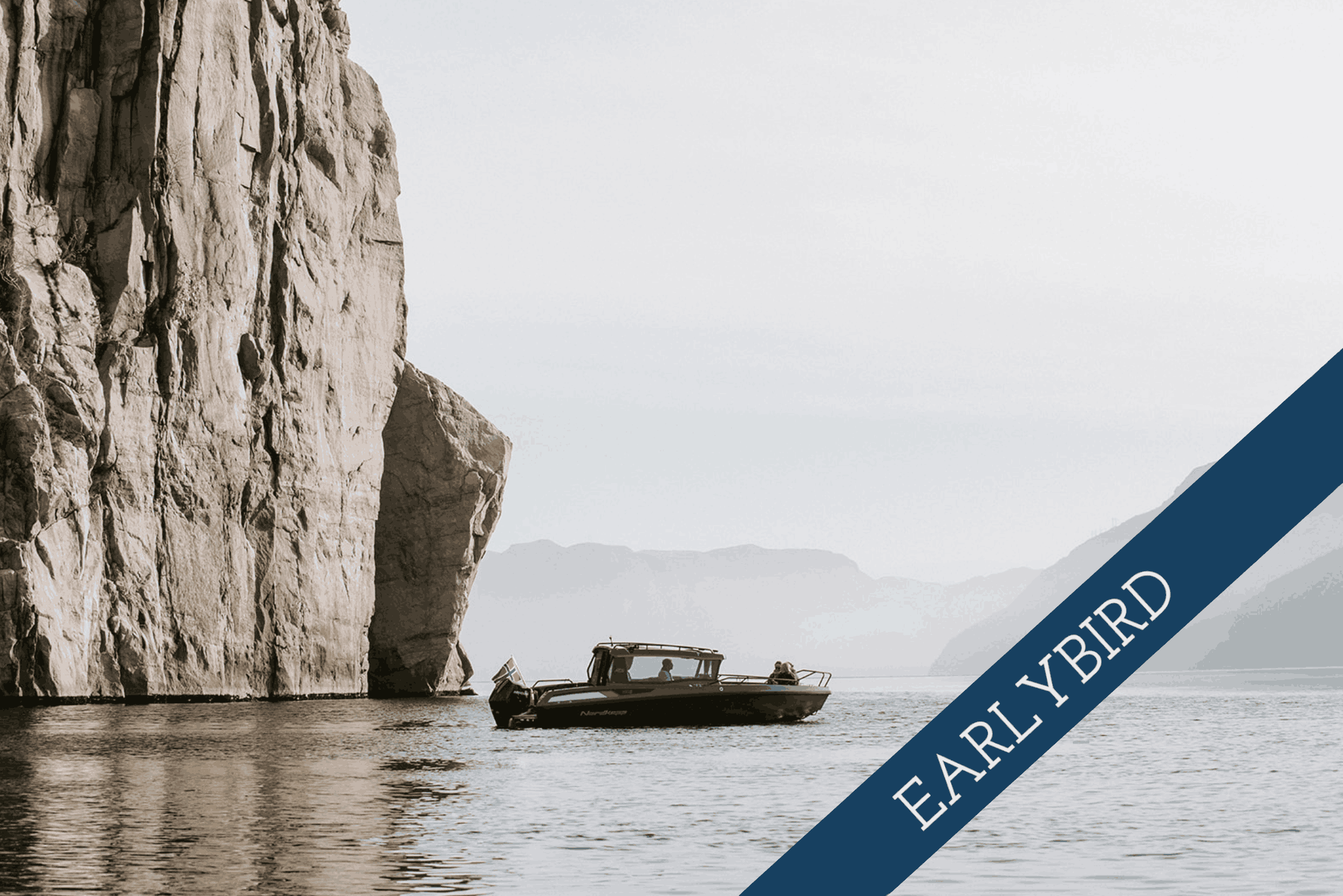 Ein Boot fährt an einer Felsklippe in einem stillen See oder Meer vorbei, im Hintergrund sind Berge sichtbar.