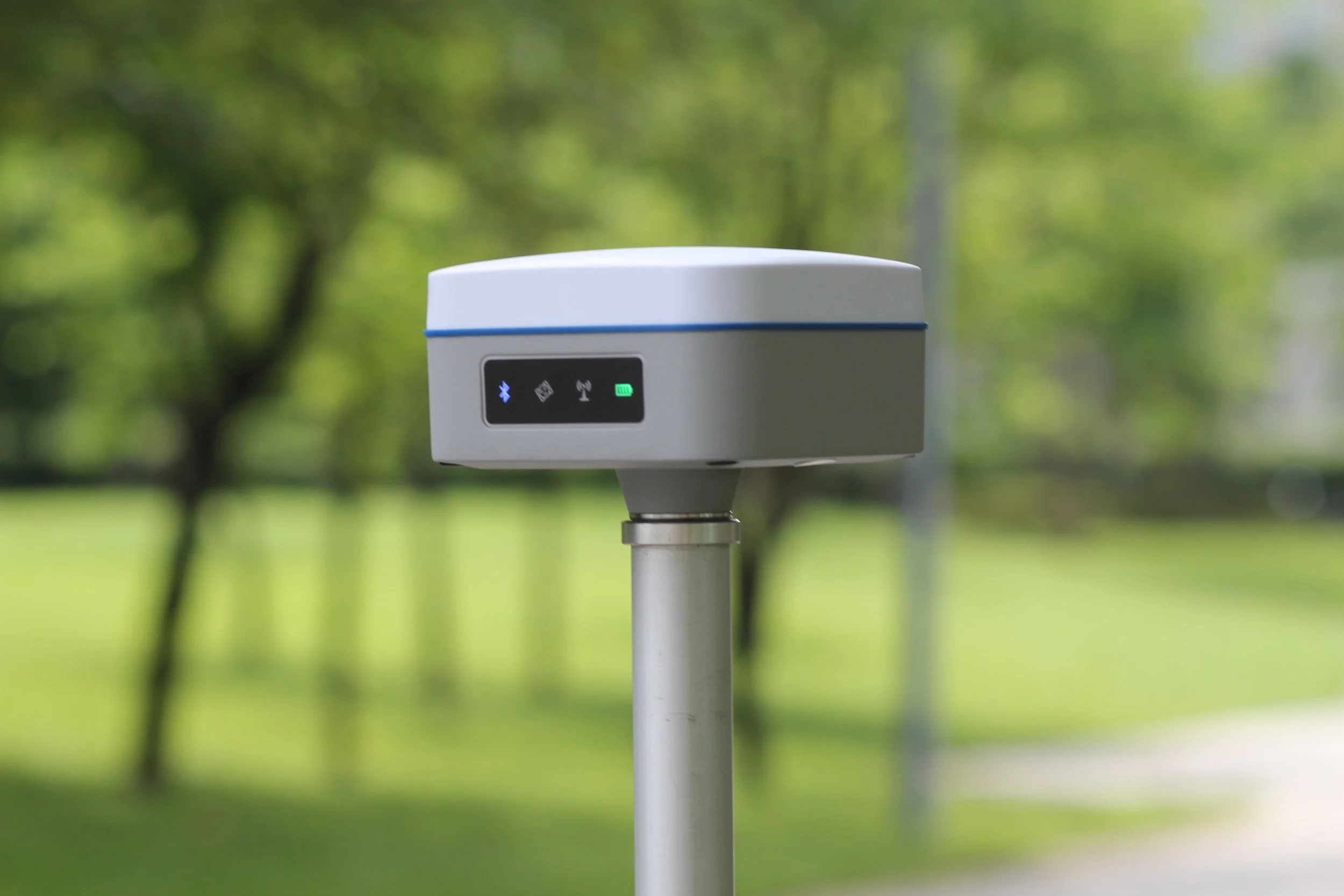 A white electronic device mounted on a pole with an LED display showing Bluetooth, NFC, signal, and battery icons, set against a green, wooded background.