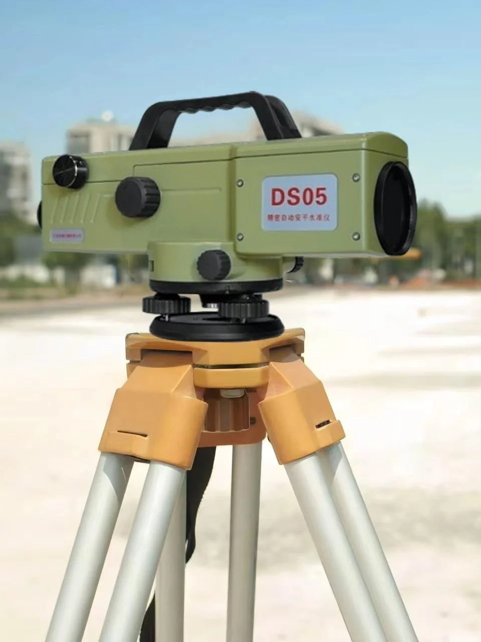 A green laser level mounted on a tripod outdoors with buildings and a blue sky in the background.