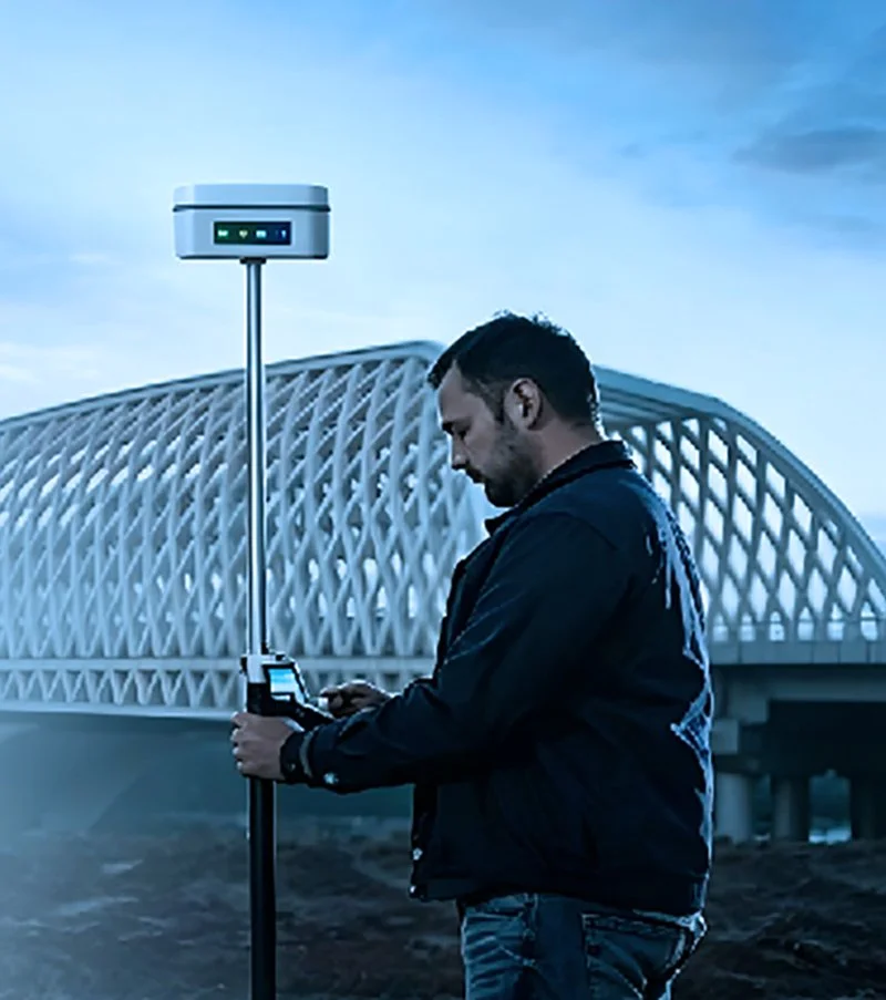 A man using a mobile device connected to a surveying or mapping device on a pole in front of a modern architectural bridge at dusk.