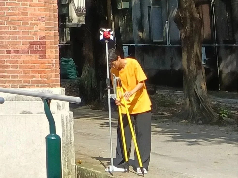 A person in a yellow shirt and dark pants using a surveying instrument outdoors near a brick building and trees.