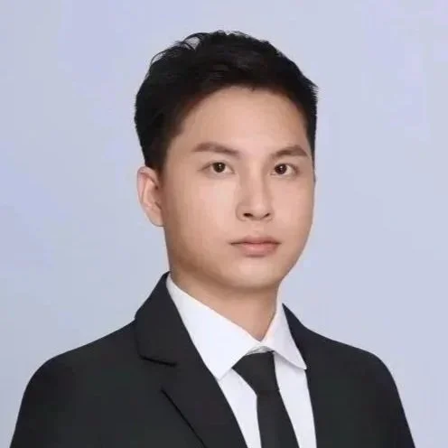 A young man with short dark hair and blue eyes taking a selfie indoors with a window behind him, showing a bright cityscape and a red Chinese decoration in the upper left corner.