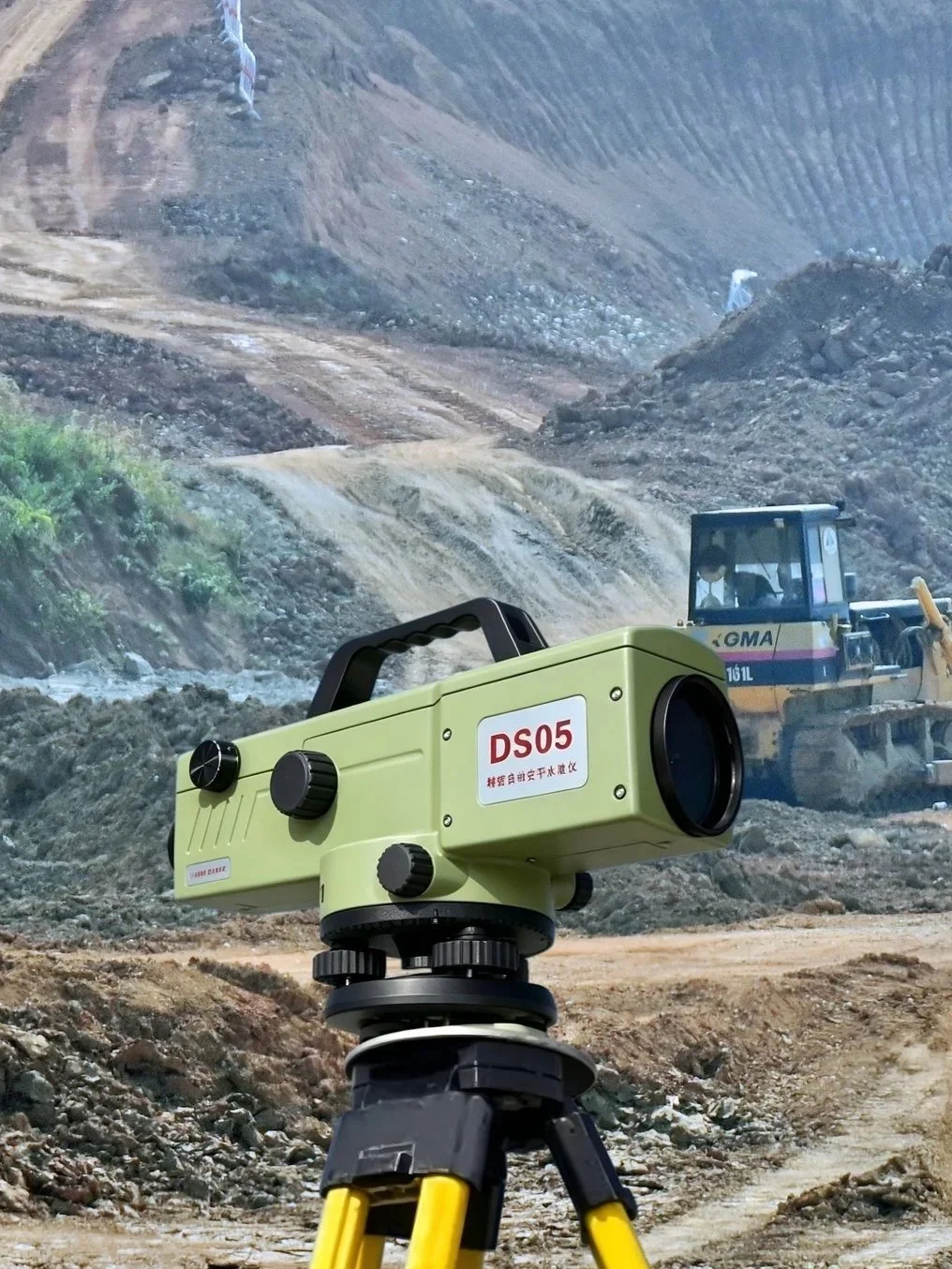 A green laser scanner on a tripod at a construction site with excavators and a dirt mountain in the background.
