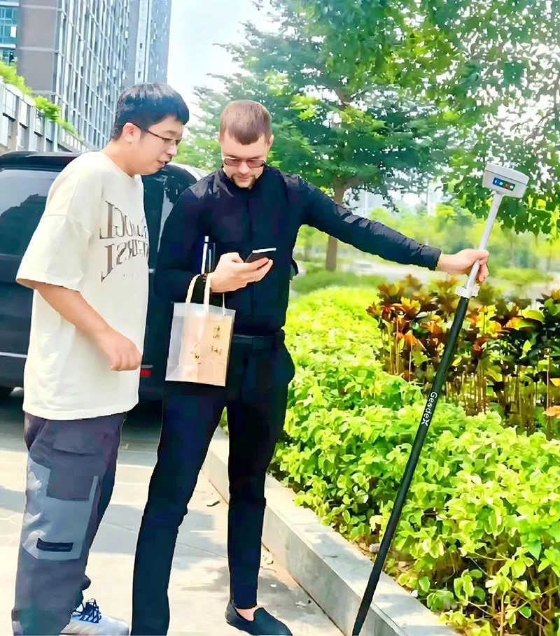 Two men standing on a sidewalk near green bushes and trees, looking at a smartphone together. One man is holding a sidewalk survey pole with a device on top, and a paper bag hangs from his arm. A black vehicle and tall buildings are visible in the background.