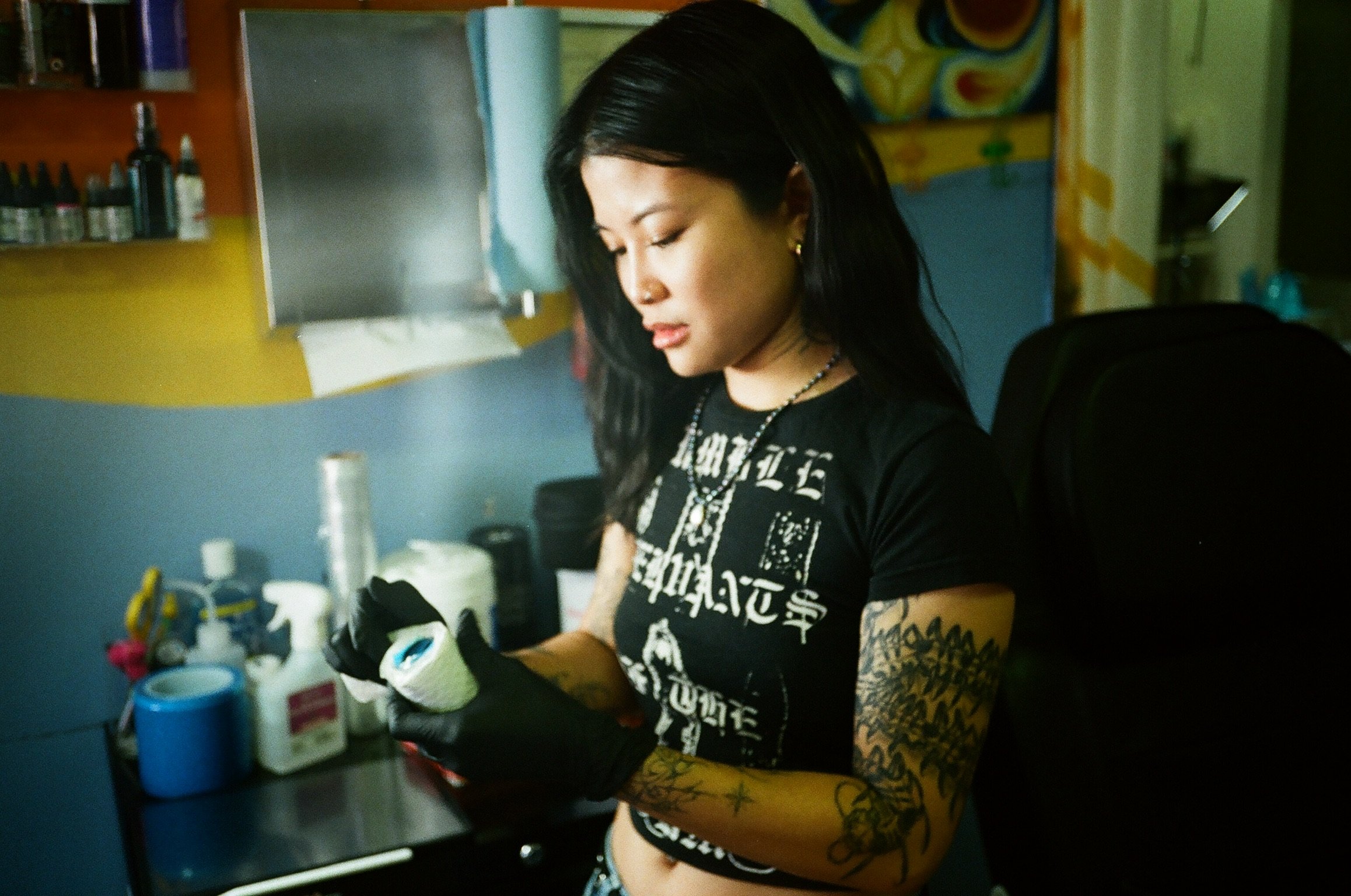 A woman with dark hair, multiple tattoos on her arms, wearing a black graphic T-shirt and gloves, is working at a tattoo station in a tattoo studio. The studio has shelves with tattoo ink bottles and other equipment in the background.