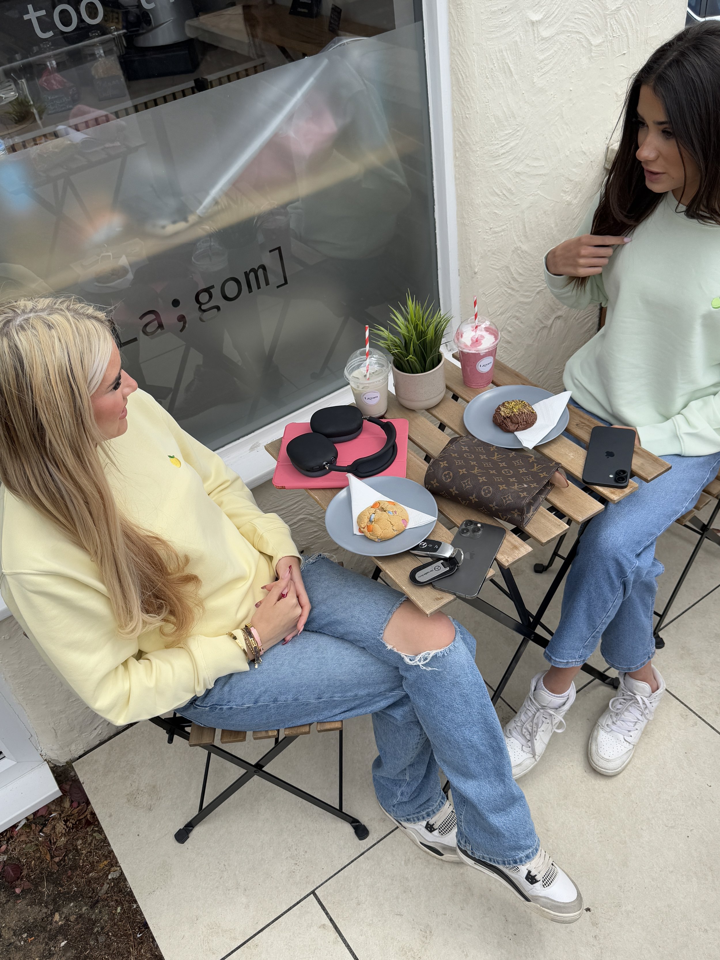 Two women sitting at a small outdoor table with drinks and snacks, engaging in conversation.