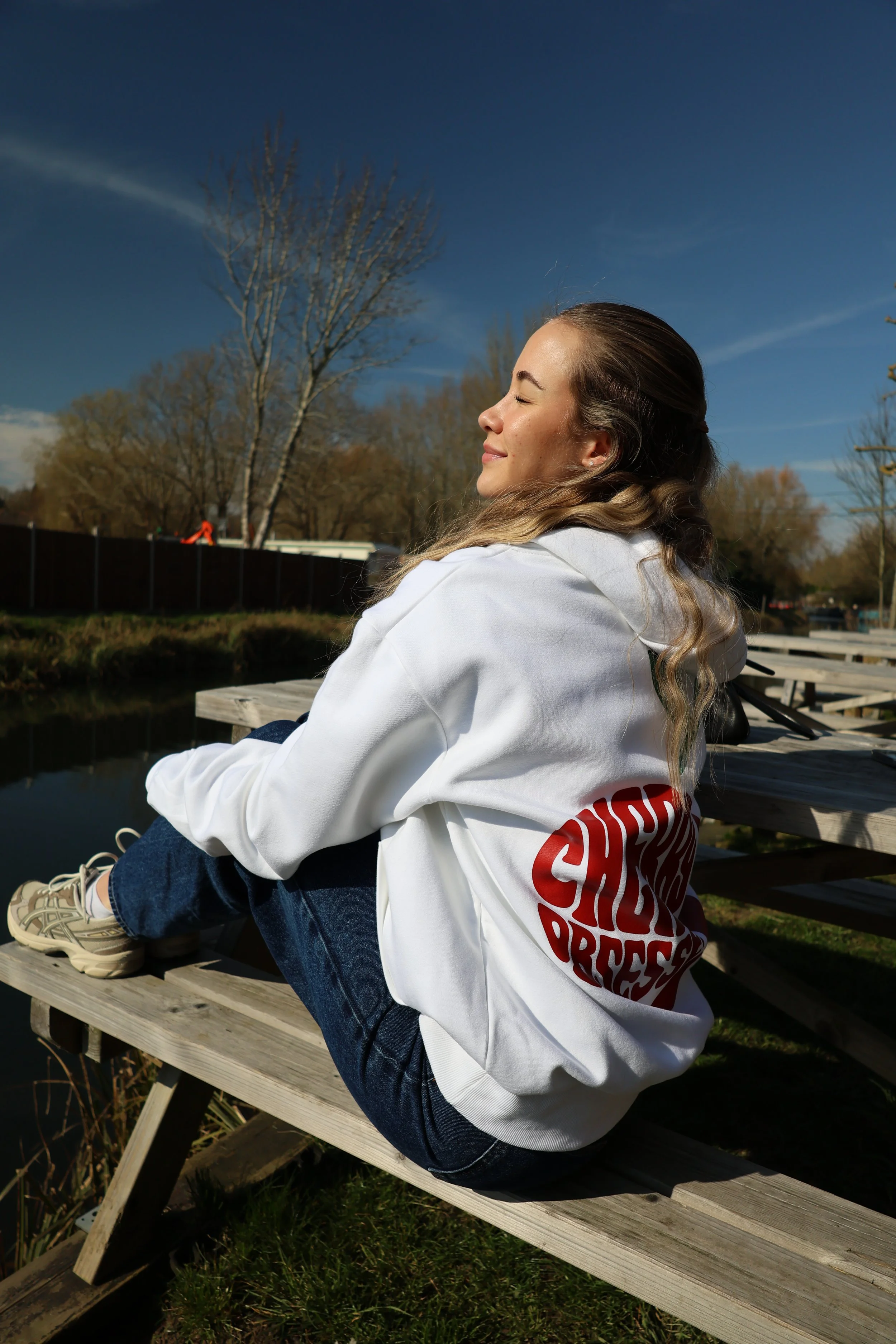 A woman sitting on a wooden bench outdoors with her eyes closed and smiling, wearing a white hoodie and jeans.