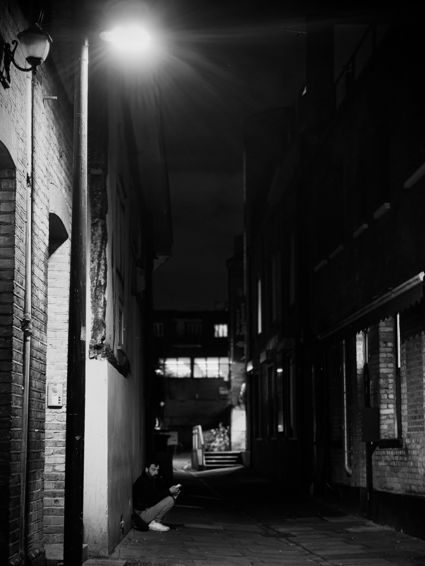 Night scene of a narrow alleyway with brick buildings on either side, illuminated by a bright streetlight, with a person sitting on the ground using a mobile phone.