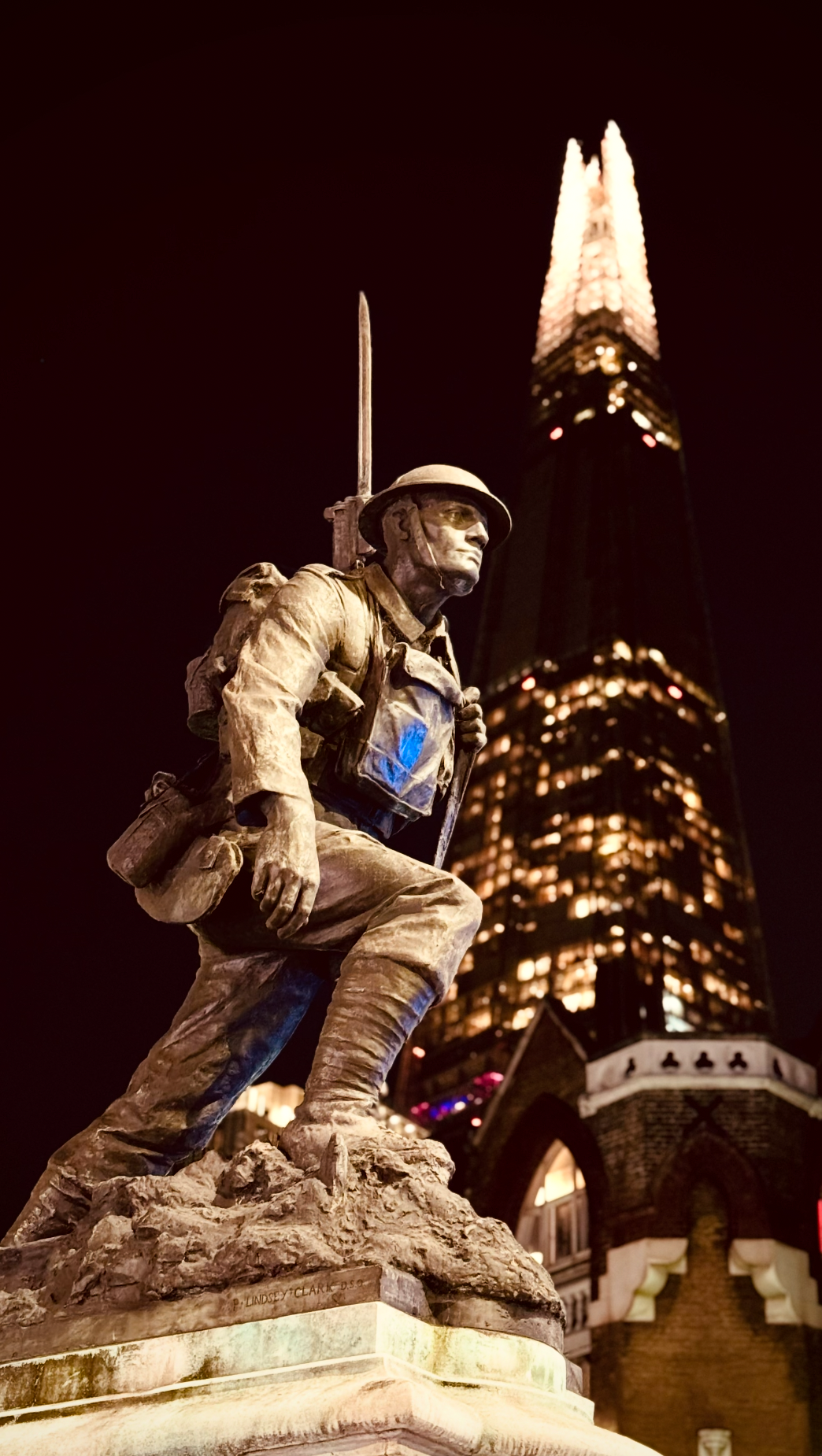 Bronze statue of a soldier with a backpack and rifle, kneeling on a rocky base, illuminated at night with the London Shard building in the background.
