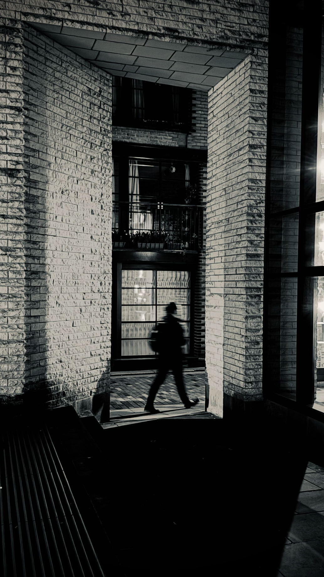 Silhouette of a person walking past a brick building at night, with illuminated windows and reflections.