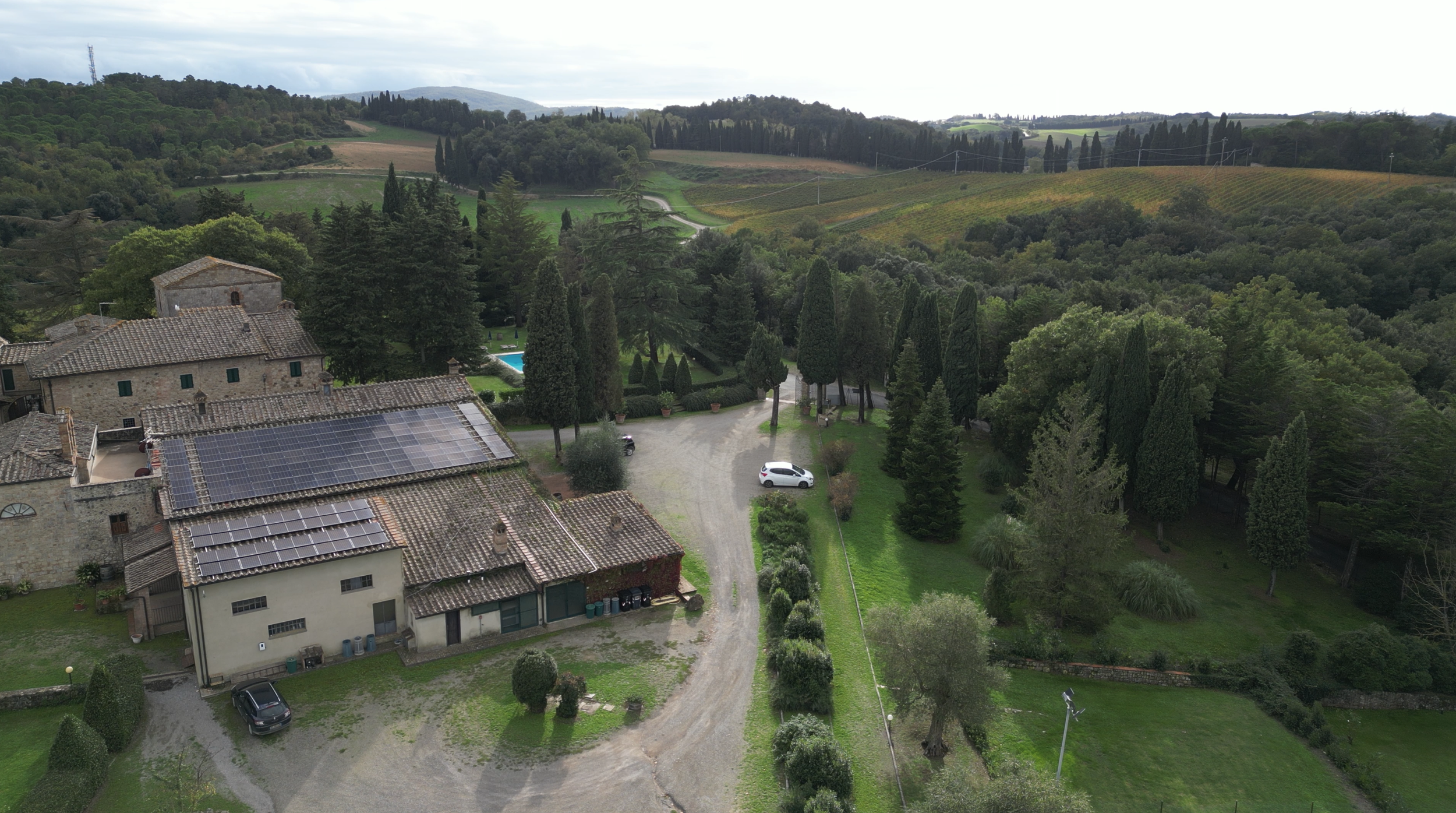 Aerial view of a countryside estate with stone buildings, some covered with solar panels, surrounded by lush greenery, tall trees, a swimming pool, and distant rolling hills and vineyards.