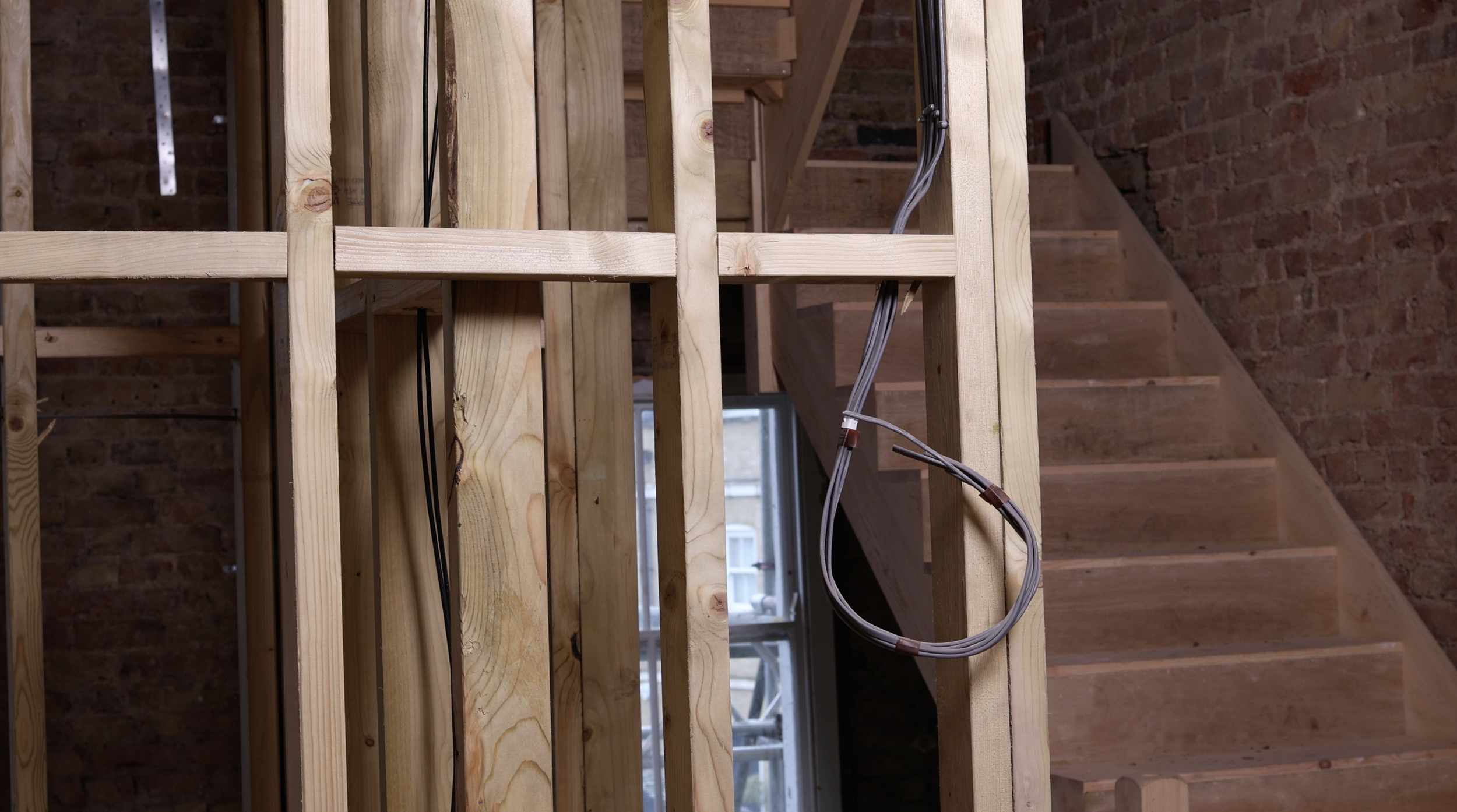 Interior of a staircase under construction with exposed wooden framing and electrical wiring, brick wall in the background, and a window.