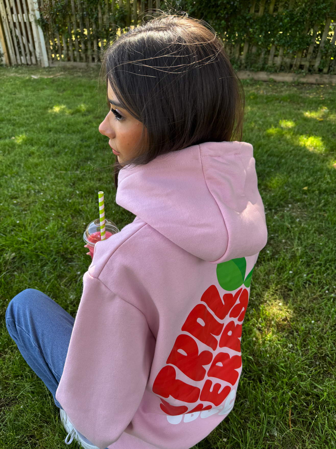 A young woman with brown hair sitting on grass in a backyard, wearing a pink hoodie with red and green graphic design on the back, holding a cup with a striped straw, with a wooden fence and green bushes in the background.