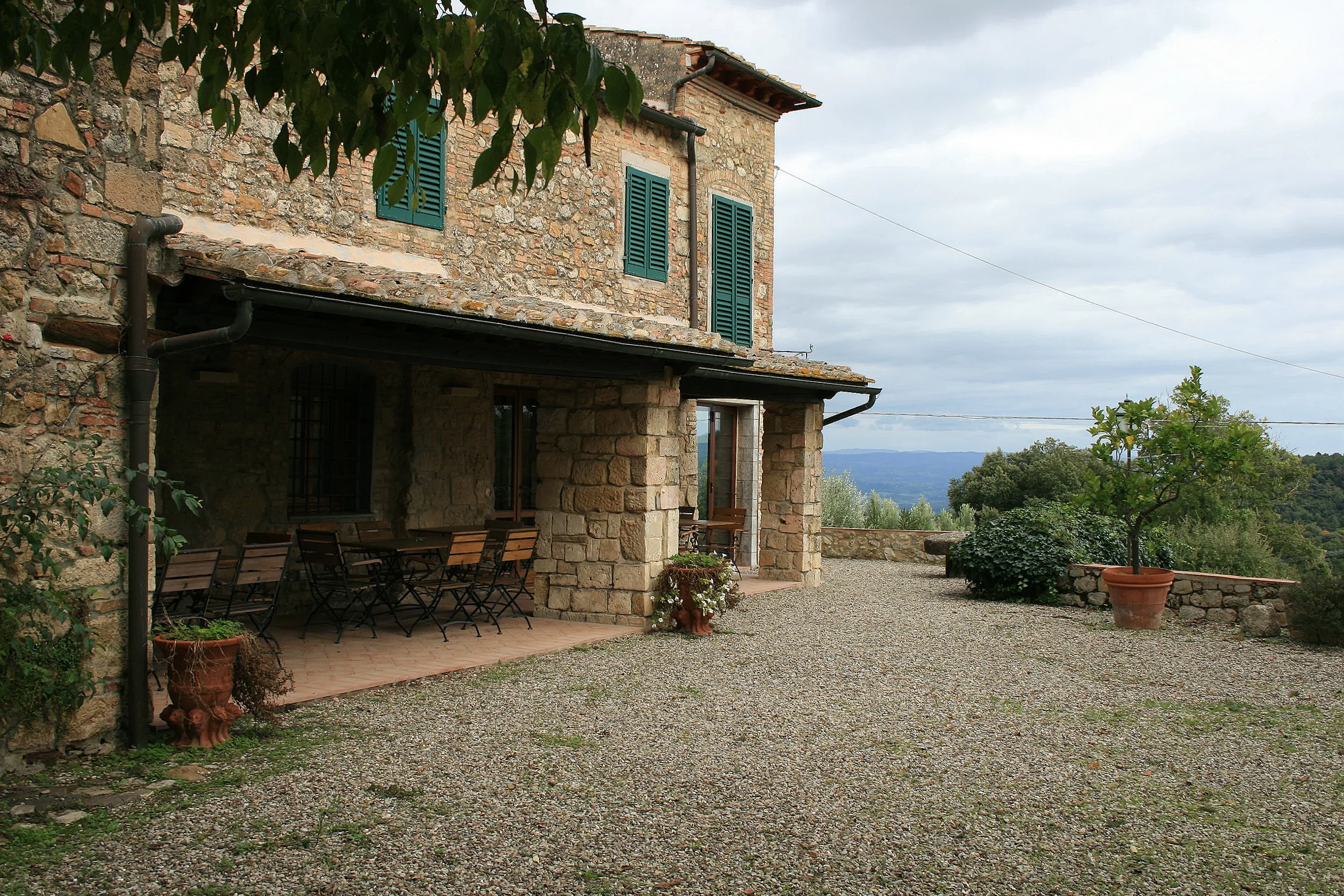 A rustic stone house with green shutters on the windows, situated on a gravel yard with a covered porch area. There are outdoor chairs, a potted plant, and trees around the house, with hills and a cloudy sky in the background.