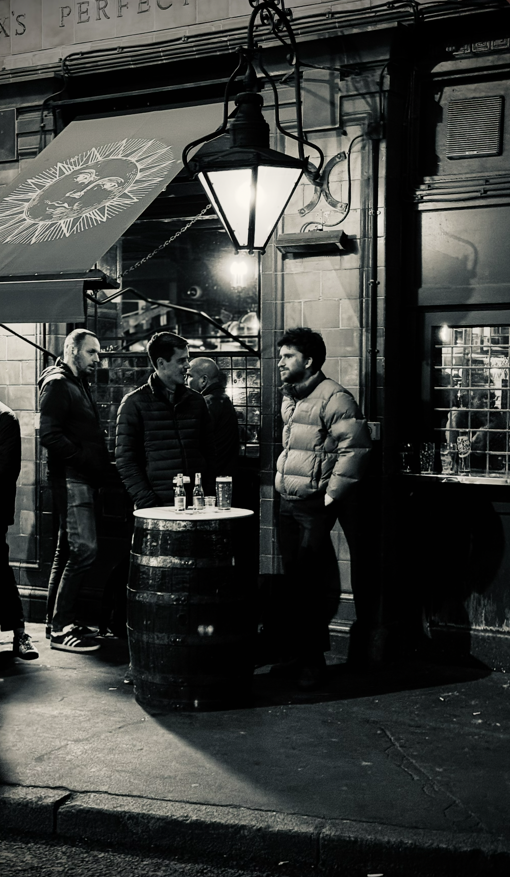 A black and white photo of four men outside a pub at night, standing near a barrel with bottles and glasses on top. They are dressed in jackets and appear to be engaged in conversation under an illuminated street lamp. The pub has a window with visib